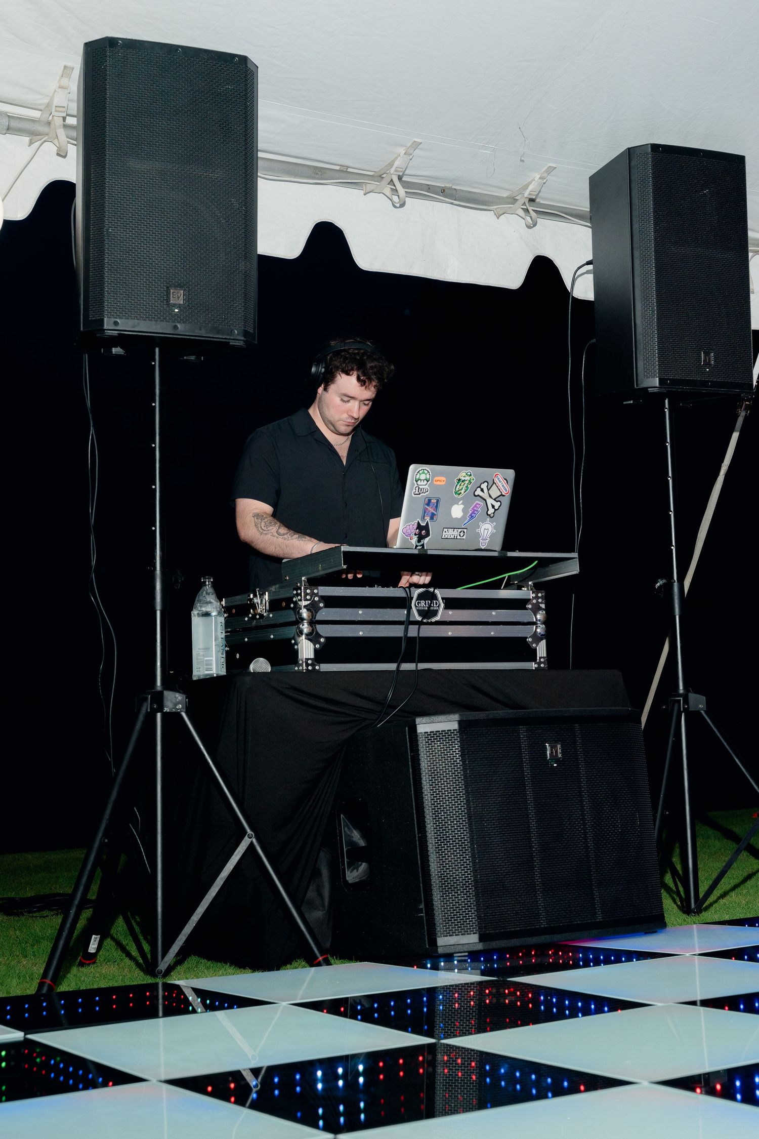 DJ in black outfit performing at outdoor event on checkered dance floor with speakers and laptop.