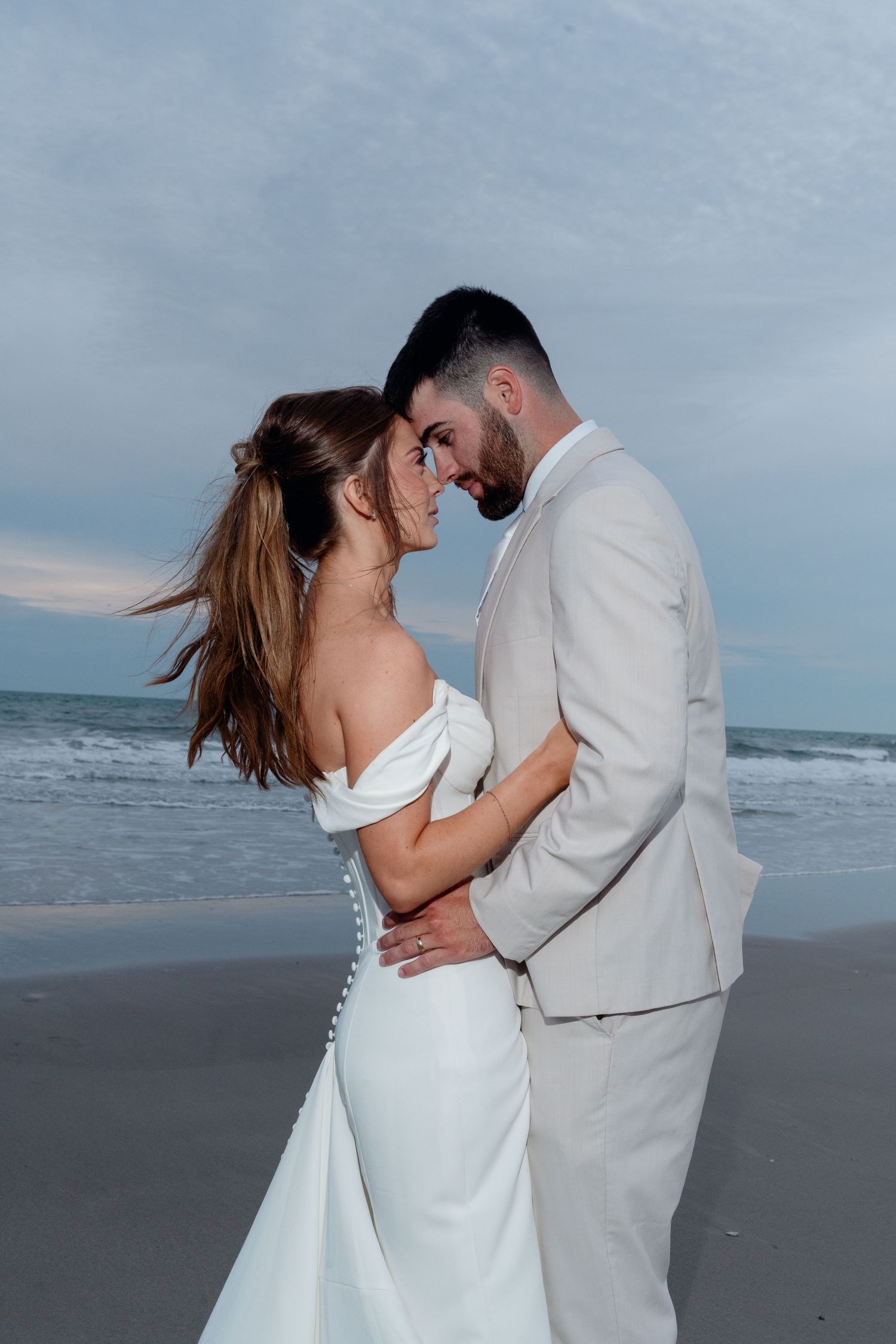 Newlywed couple sharing romantic moment on beach at dusk, bride in off-shoulder gown and groom in tan suit.