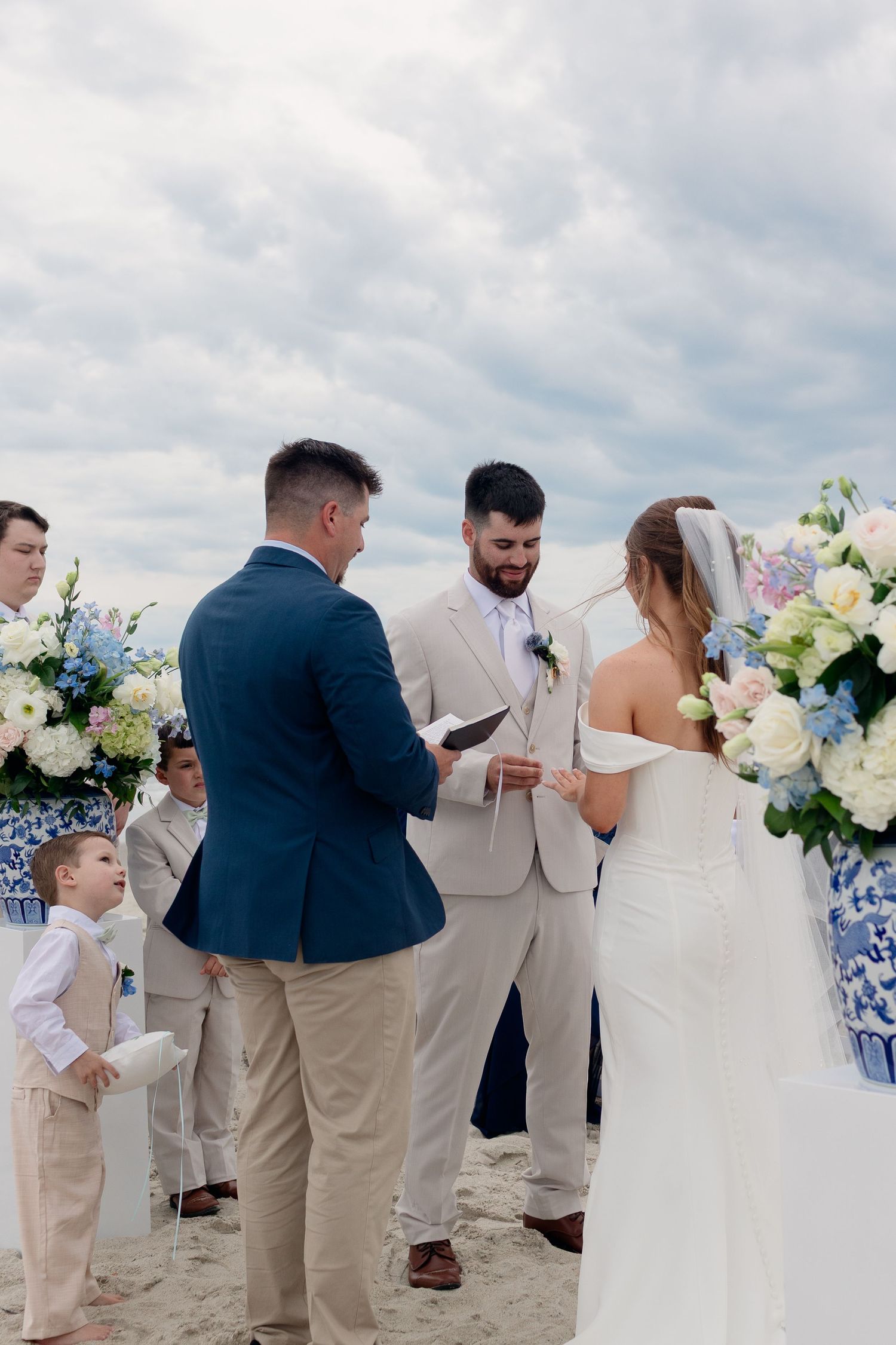 Beach wedding ceremony with groom reading vows, officiant, and bride surrounded by blue and white floral arrangements.