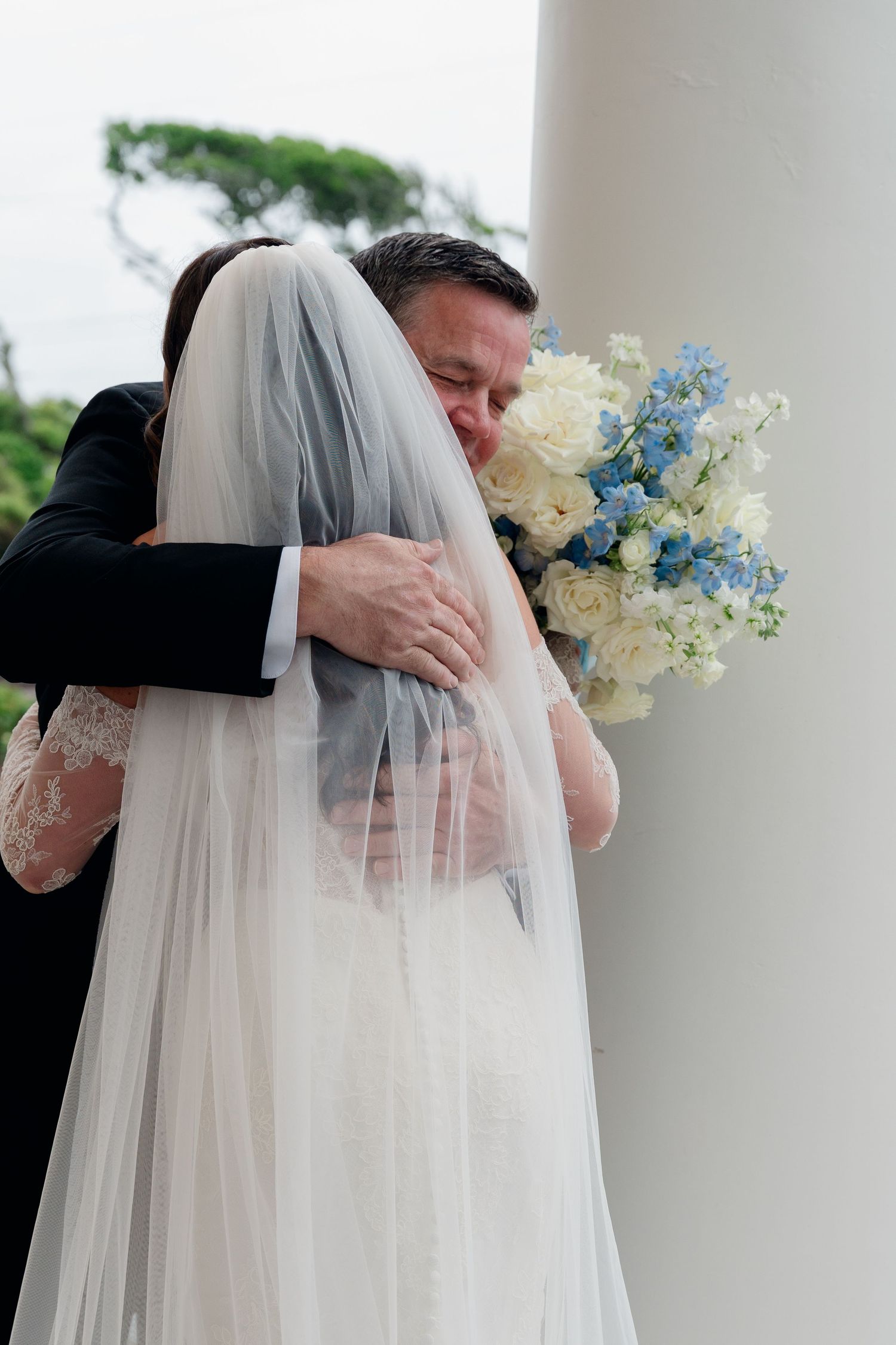Emotional father hugs bride in white lace gown holding blue and white bouquet near white column at wedding venue.