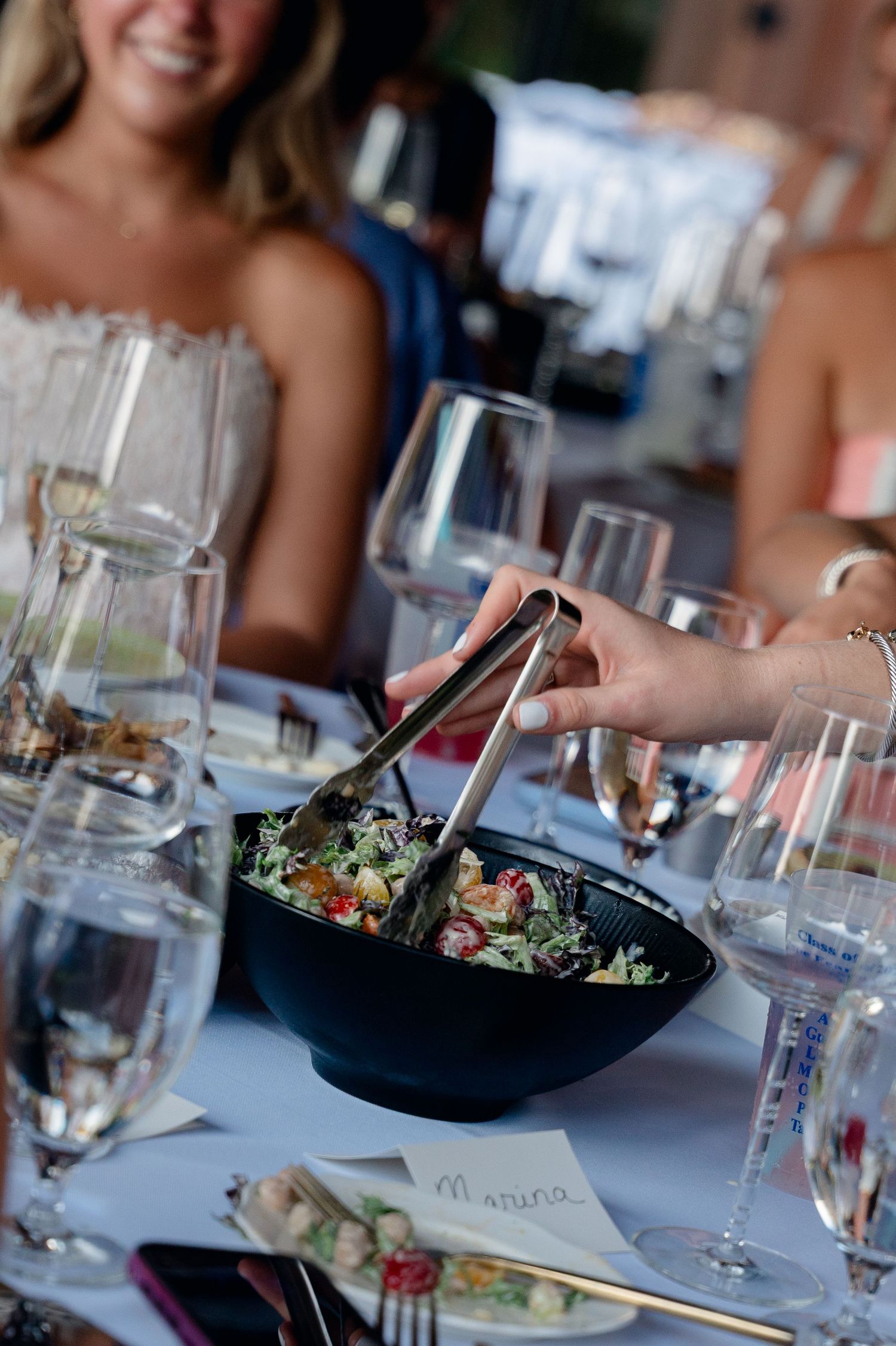 Hand serving fresh garden salad with tongs at elegantly set dining table with wine glasses.