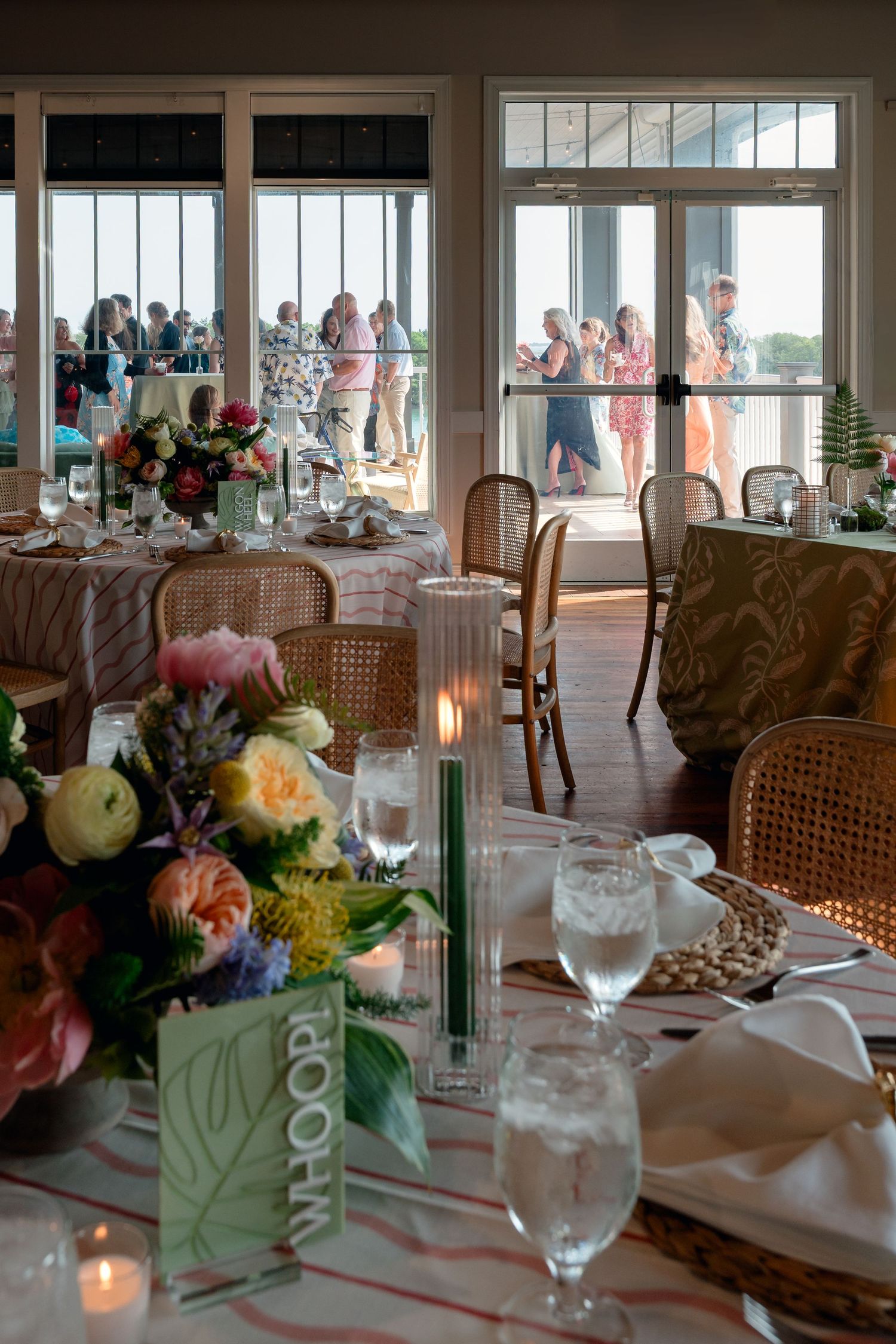 Elegant waterfront event venue with floral centerpieces, candlelit tables, and guests mingling on an outdoor deck.