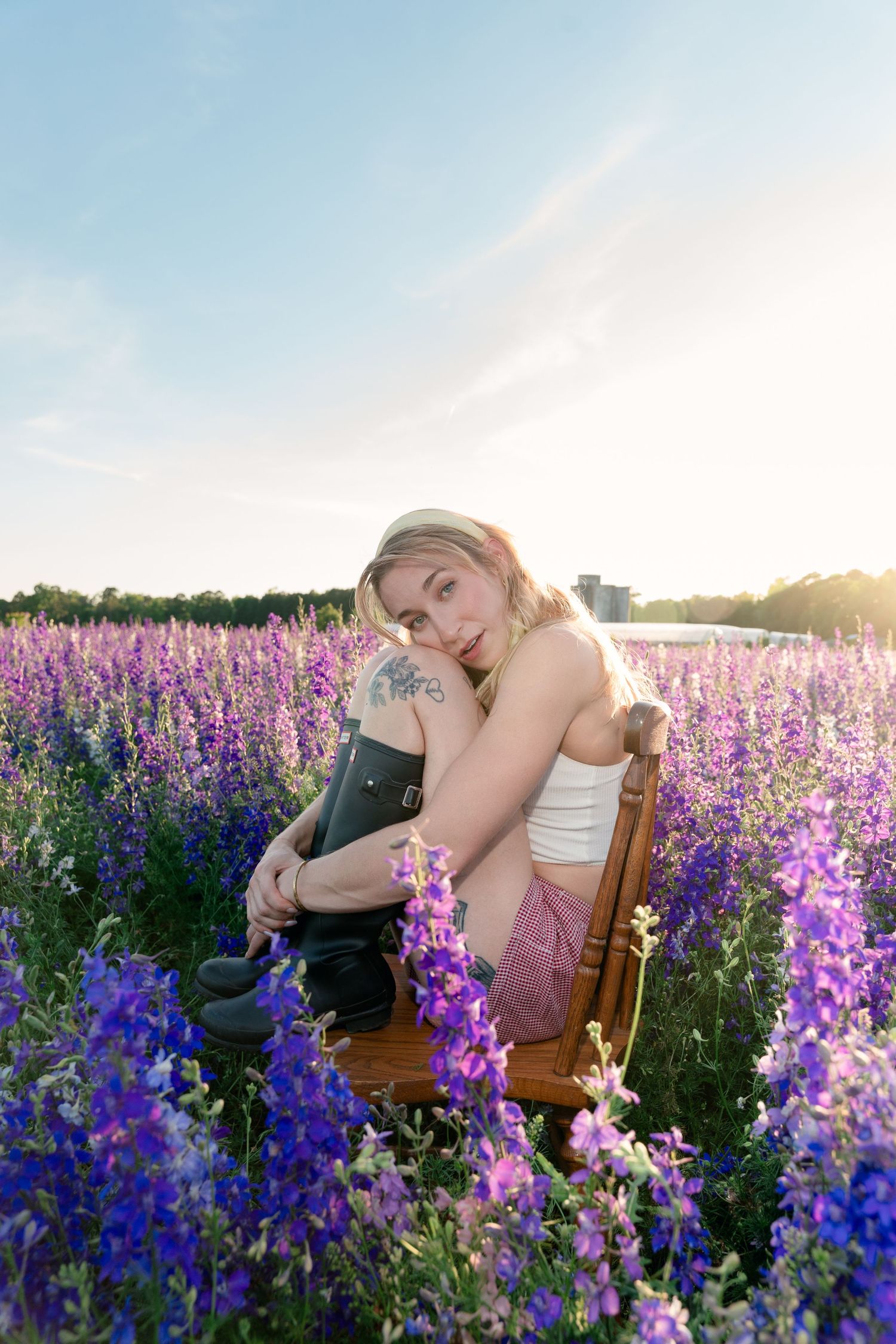 Tattooed blonde woman in white tank and shorts sitting in a purple lupine flower field at sunset.