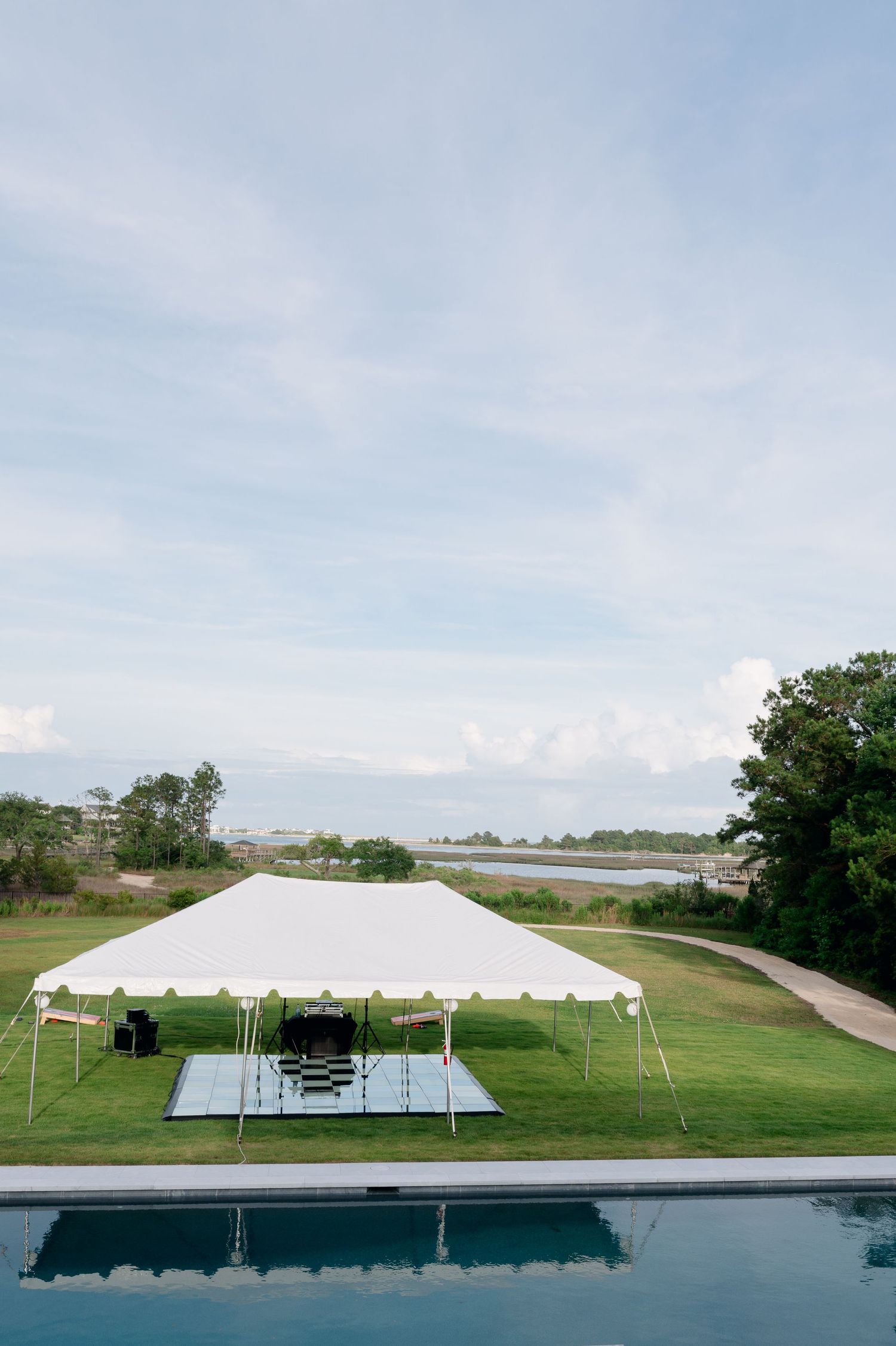 White event tent with black and white dance floor set on green lawn beside pool with water view.