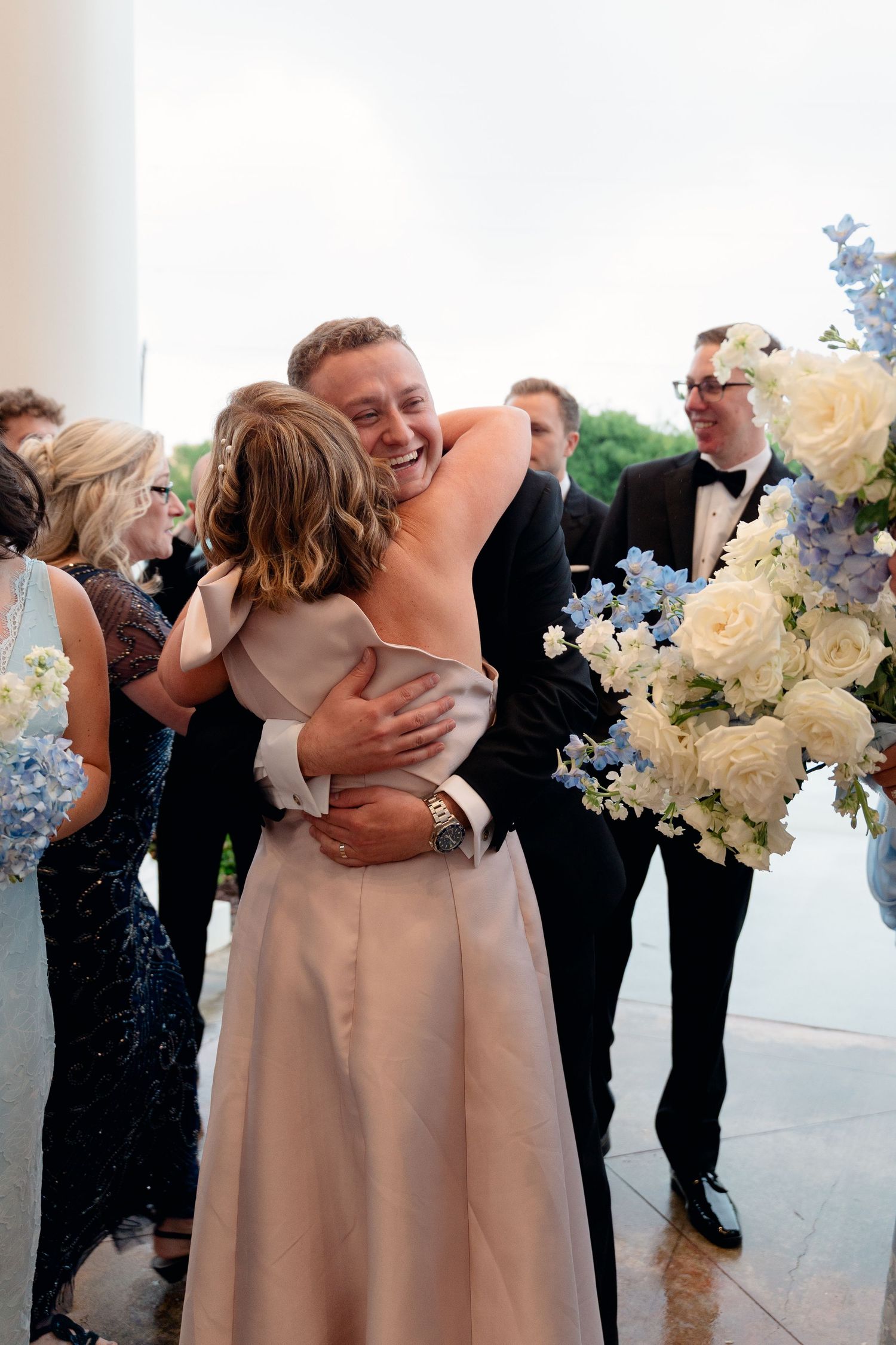Smiling groom in tuxedo embraces guest at wedding reception while bridesmaids hold blue and white floral bouquets nearby.