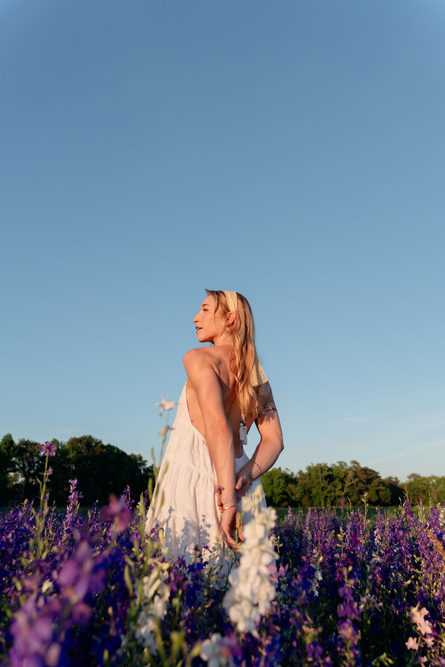 Blonde woman in white sundress standing in purple wildflower field under clear blue sky at golden hour.
