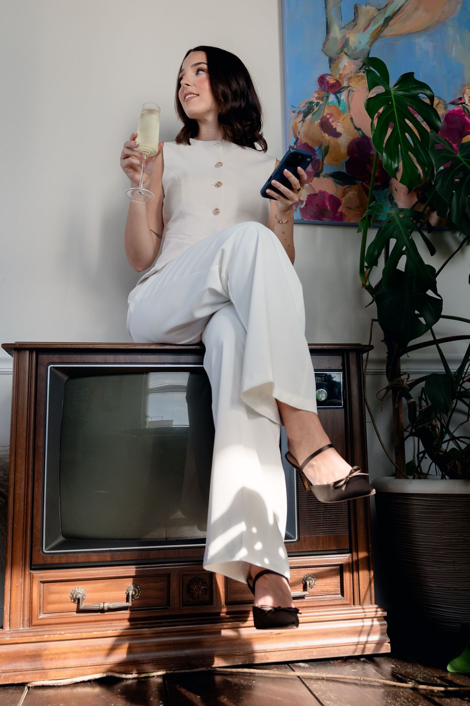 Stylish woman in white wide-leg pants sitting on vintage TV holding champagne and smartphone.