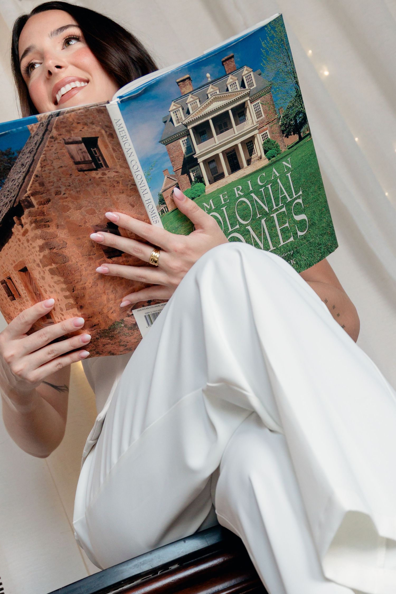 Smiling brunette woman reading American Colonial Homes book wearing white wide-leg pants indoors.