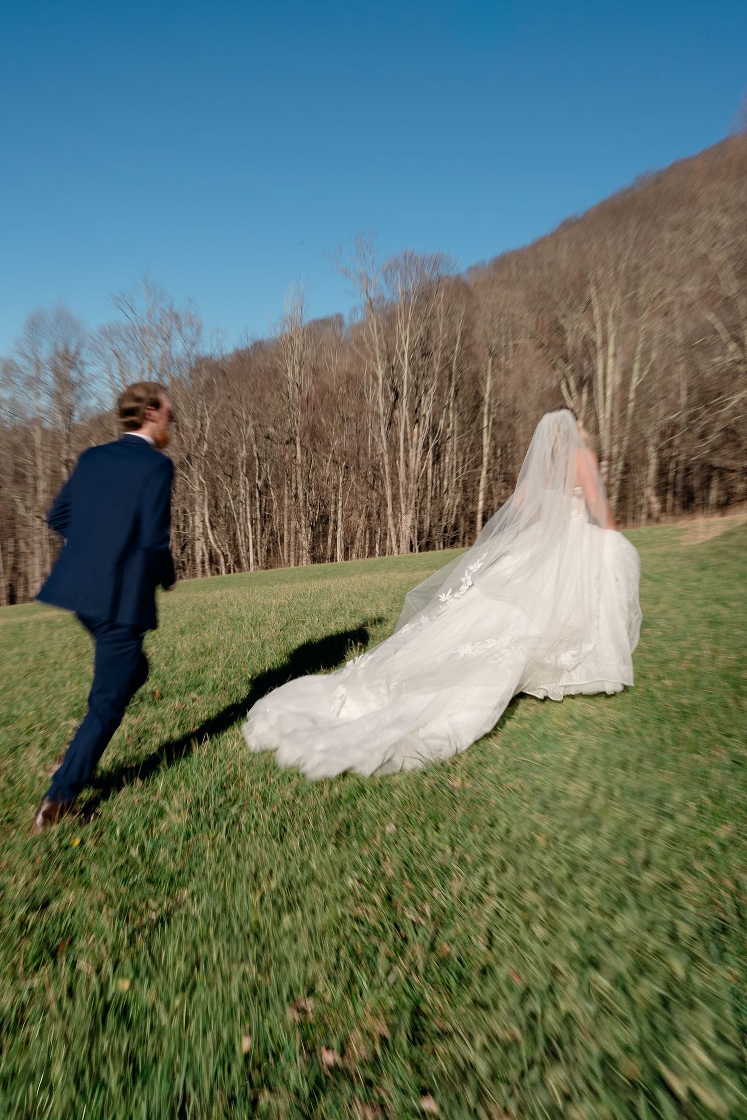 Bride in flowing white gown and veil walks with groom in navy suit across green field with bare winter trees.