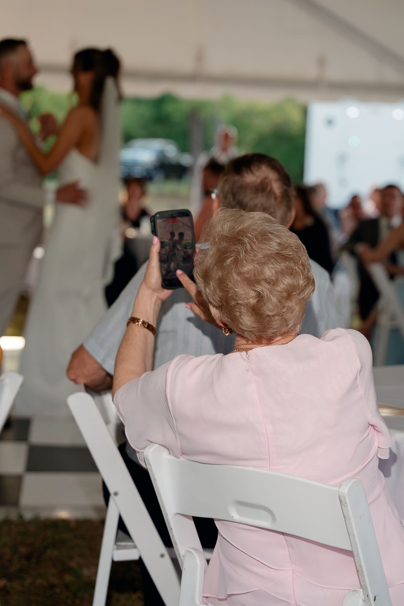 Elderly wedding guest in pink photographing the bride and groom's first dance on her smartphone.