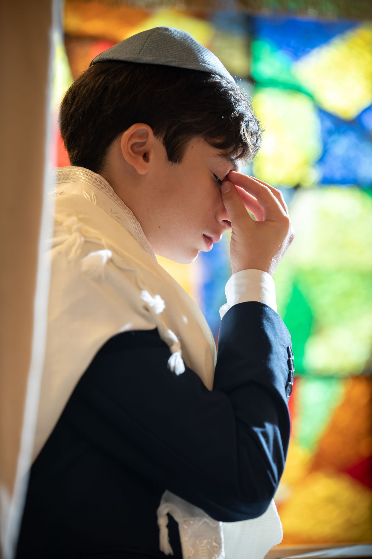 Young Jewish boy in tallit and kippah praying solemnly during his Bar Mitzvah ceremony near stained glass window.