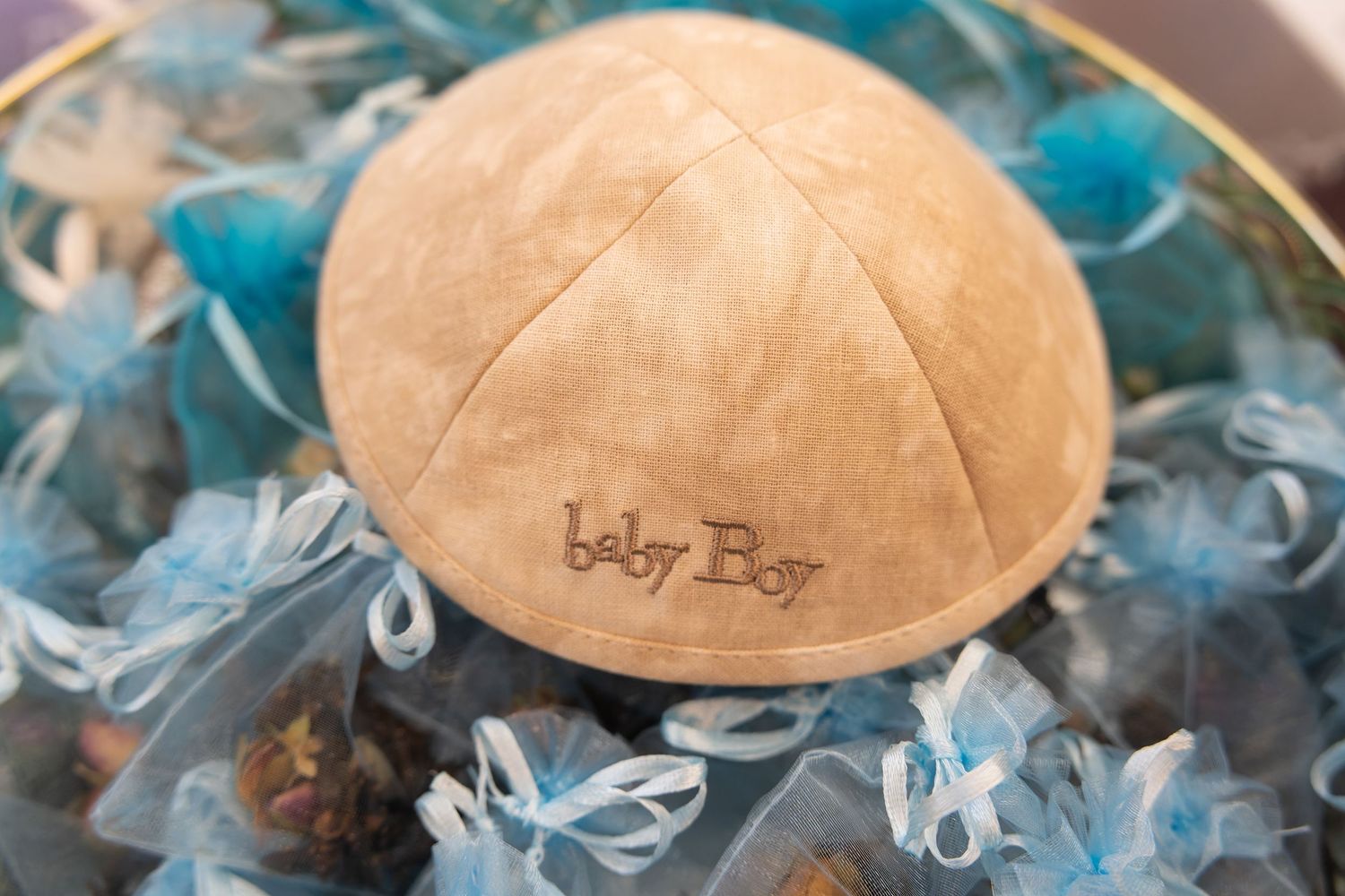 A tan suede kippah embroidered with Baby Boy text, surrounded by blue organza favor bags for a Jewish baby shower.