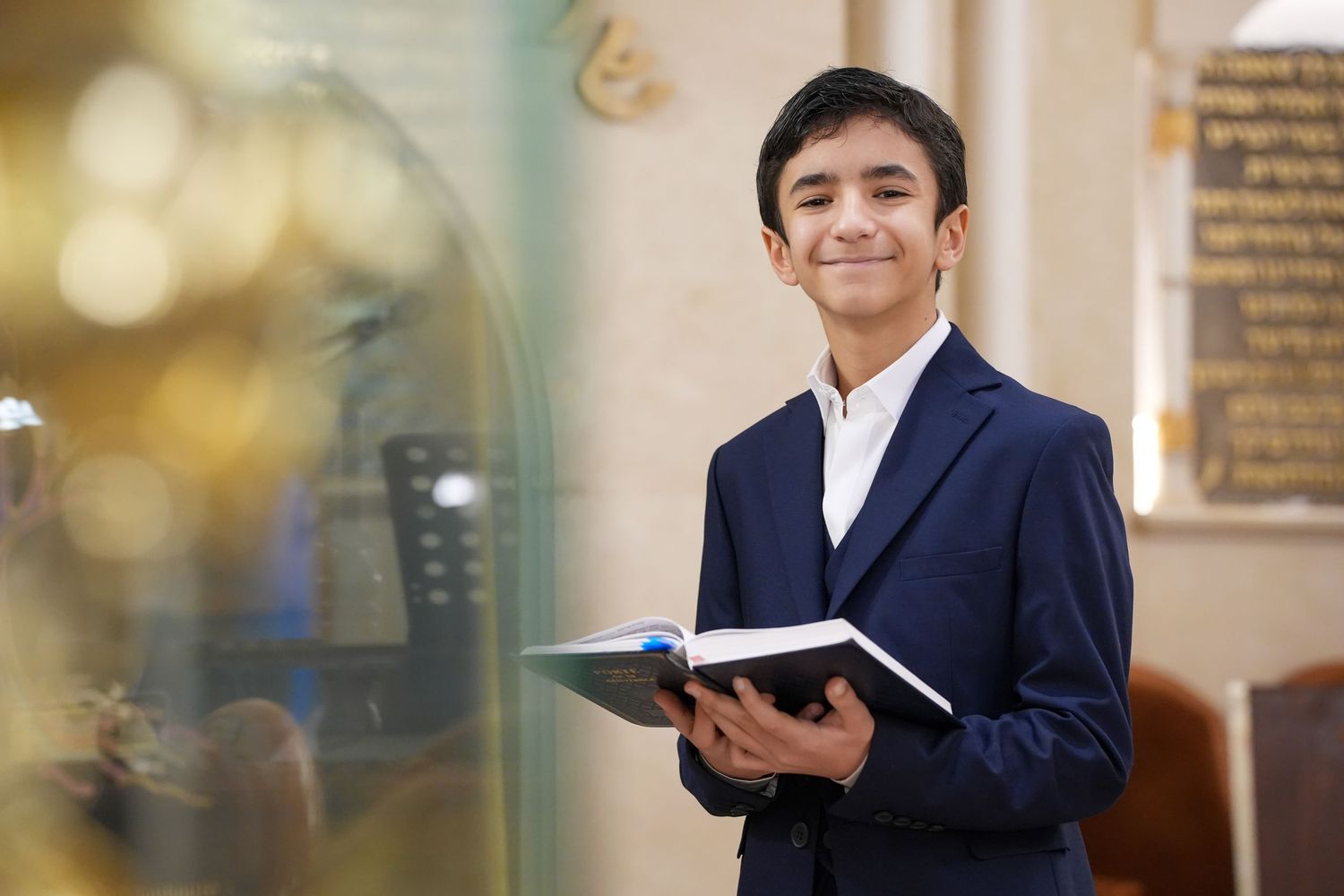 Smiling teenage boy in blue suit holds prayer book inside synagogue.