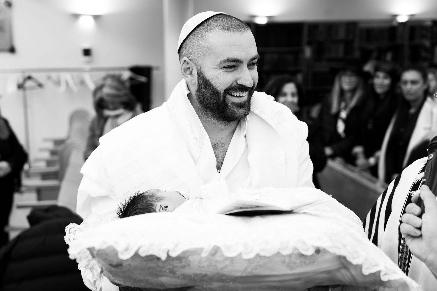 Man in white robe and kippah smiling while holding a newborn baby during a Jewish bris ceremony.