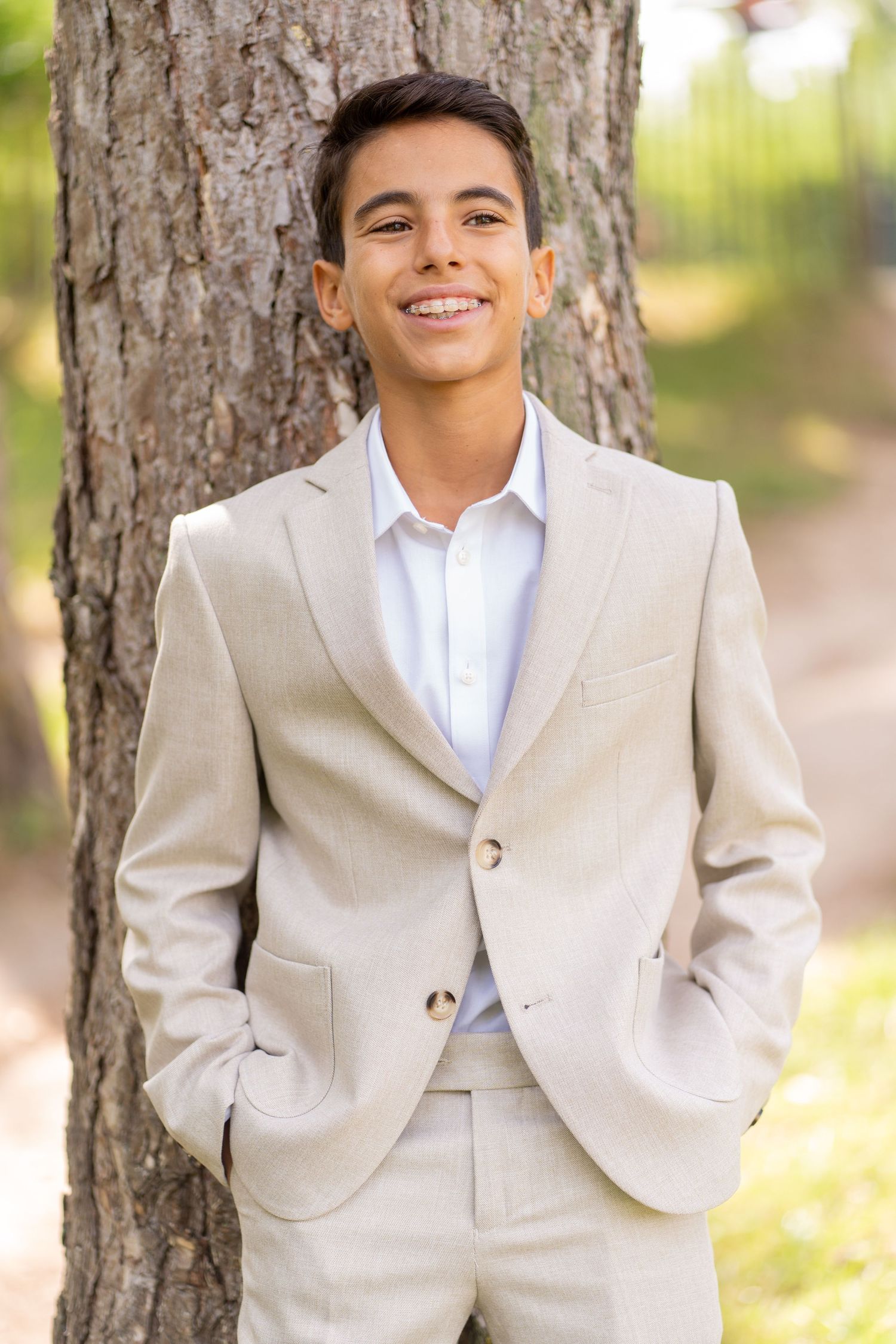 Happy teenage boy in beige two-button suit standing confidently against tree in park.