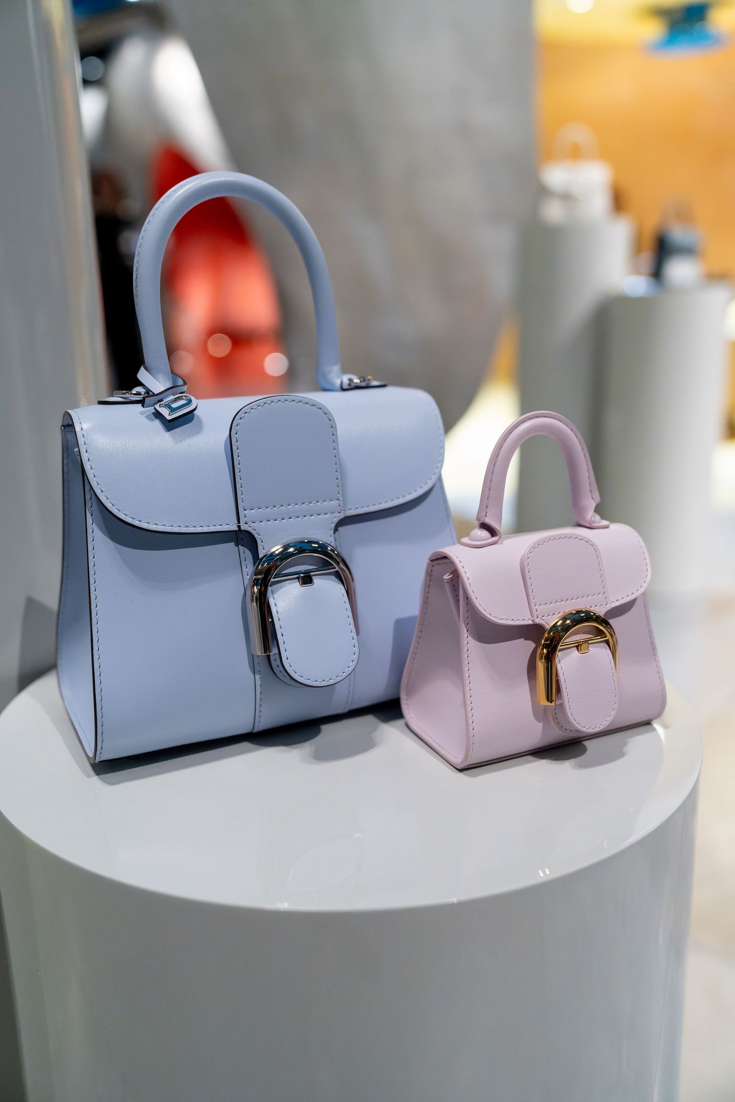 Two structured leather handbags in pastel blue and pink displayed on white pedestals in a luxury boutique.