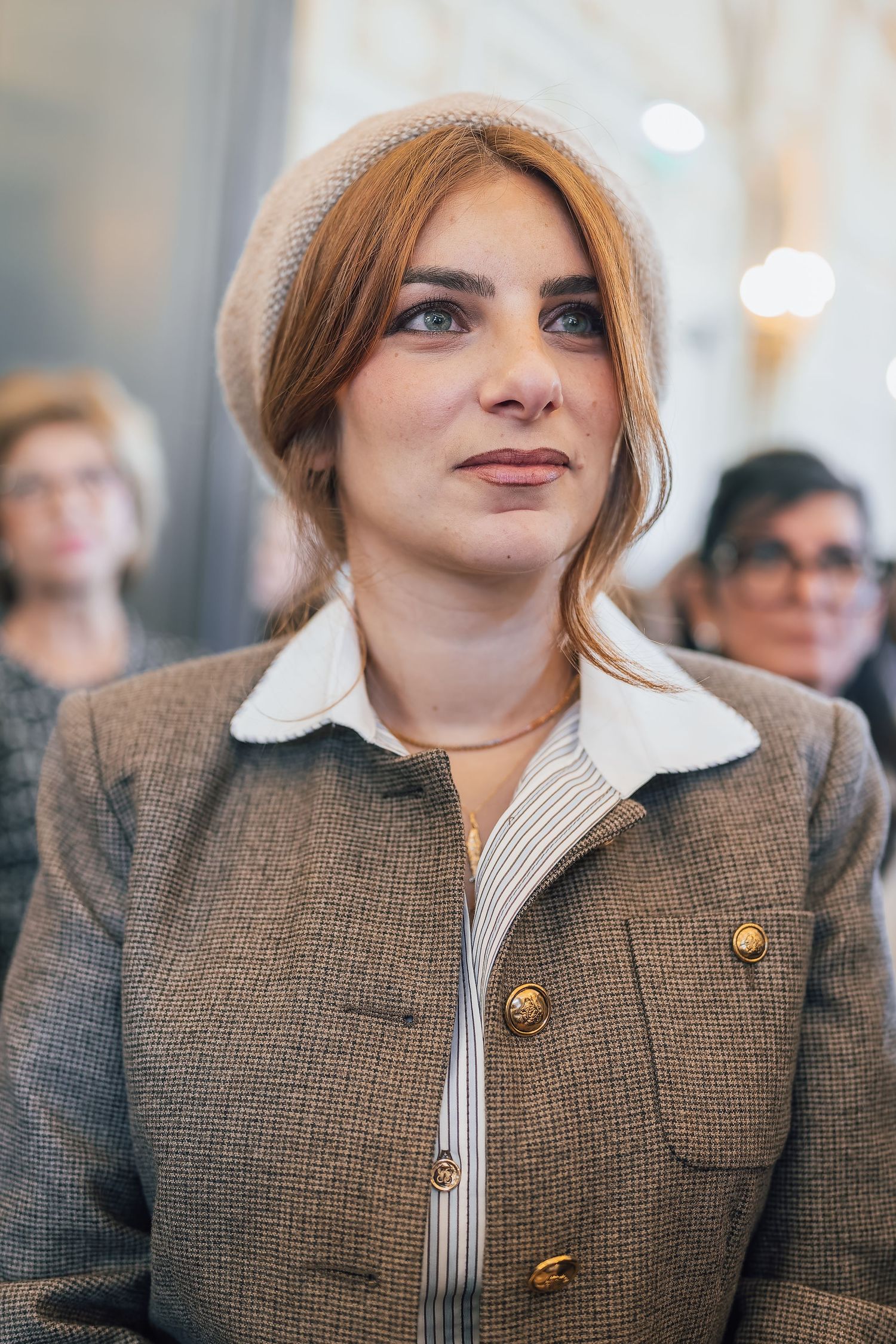 Young woman in beige beret and tweed blazer with gold buttons, posing indoors at a formal event.
