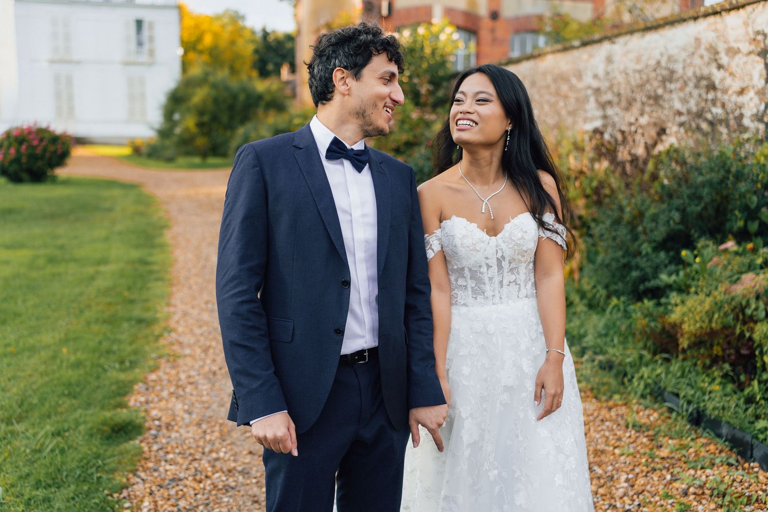 Happy newlyweds walking hand in hand on a garden path, groom in navy tuxedo, bride in off-shoulder lace wedding dress.