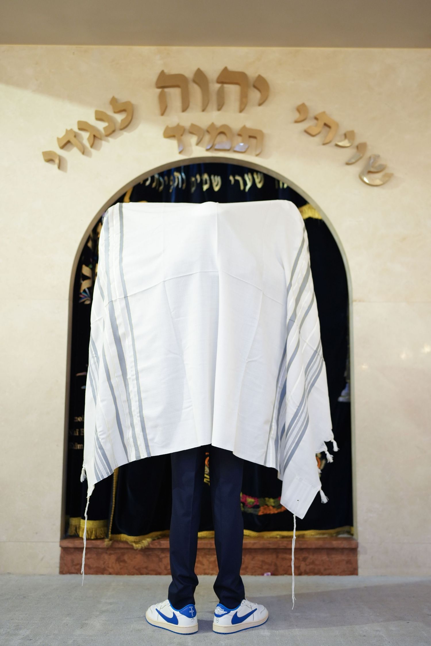 Boy wrapped in white tallit prayer shawl stands before Hebrew inscription in synagogue.