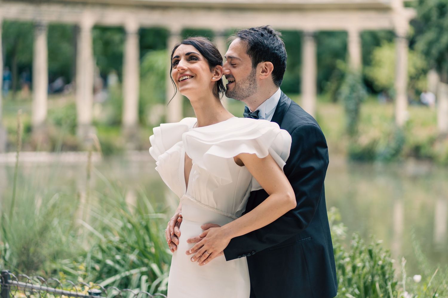 Bride in ruffled white gown embraced by groom in dark suit near scenic garden pond.