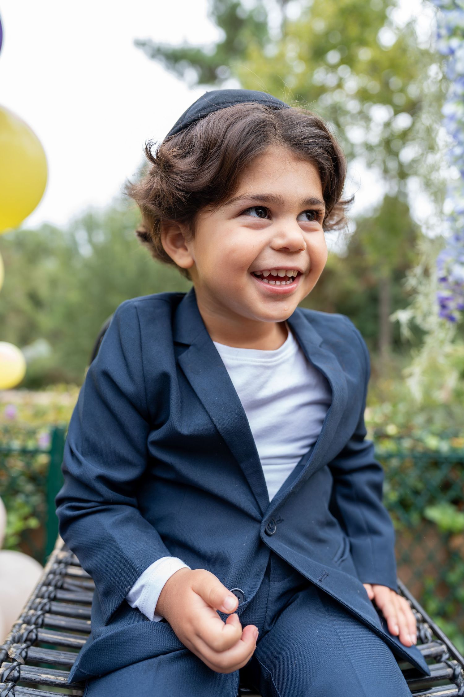 Happy young boy in navy suit and kippah laughing at an outdoor Jewish celebration with balloons.