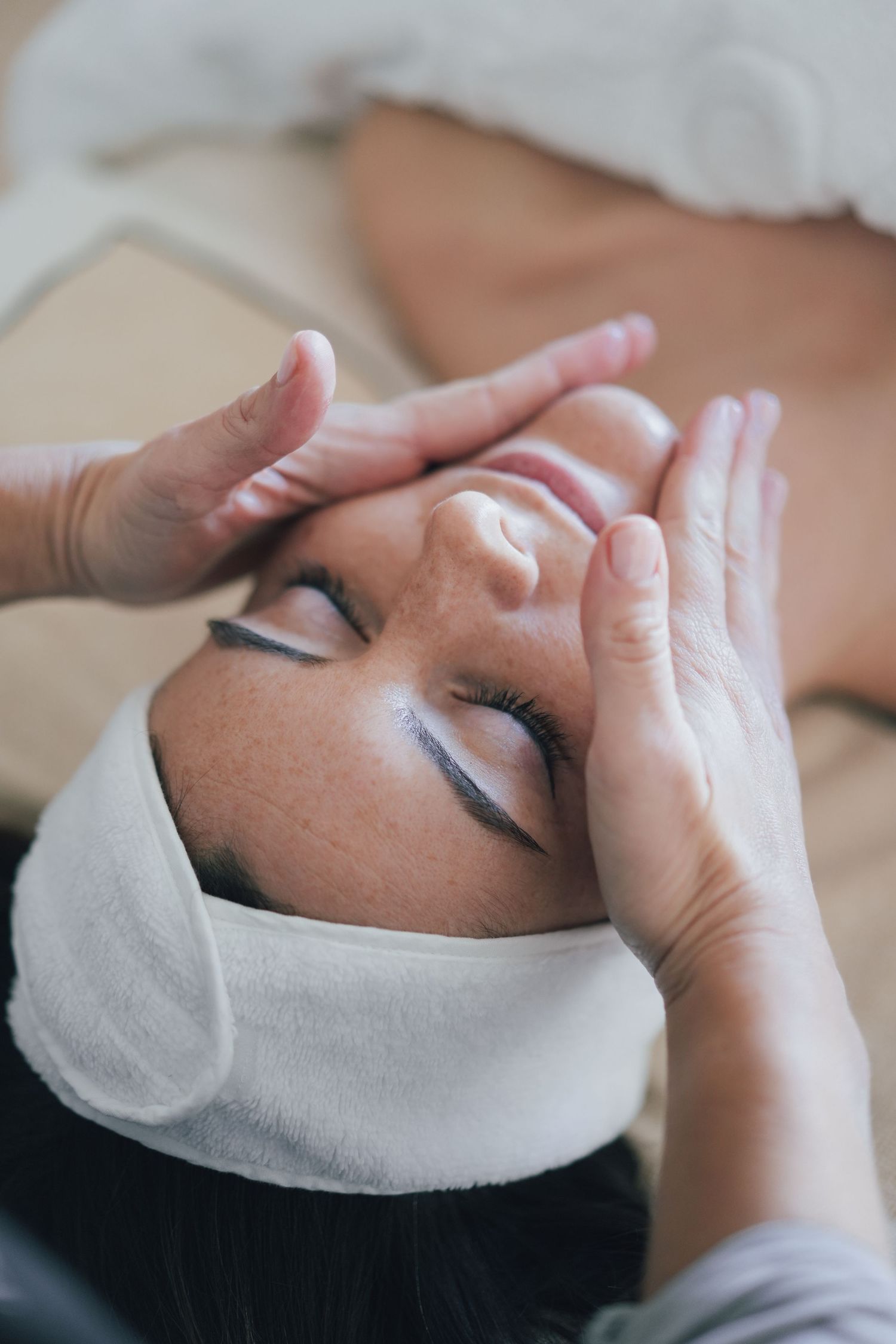 Esthetician performing relaxing facial massage on woman with white headband at spa.