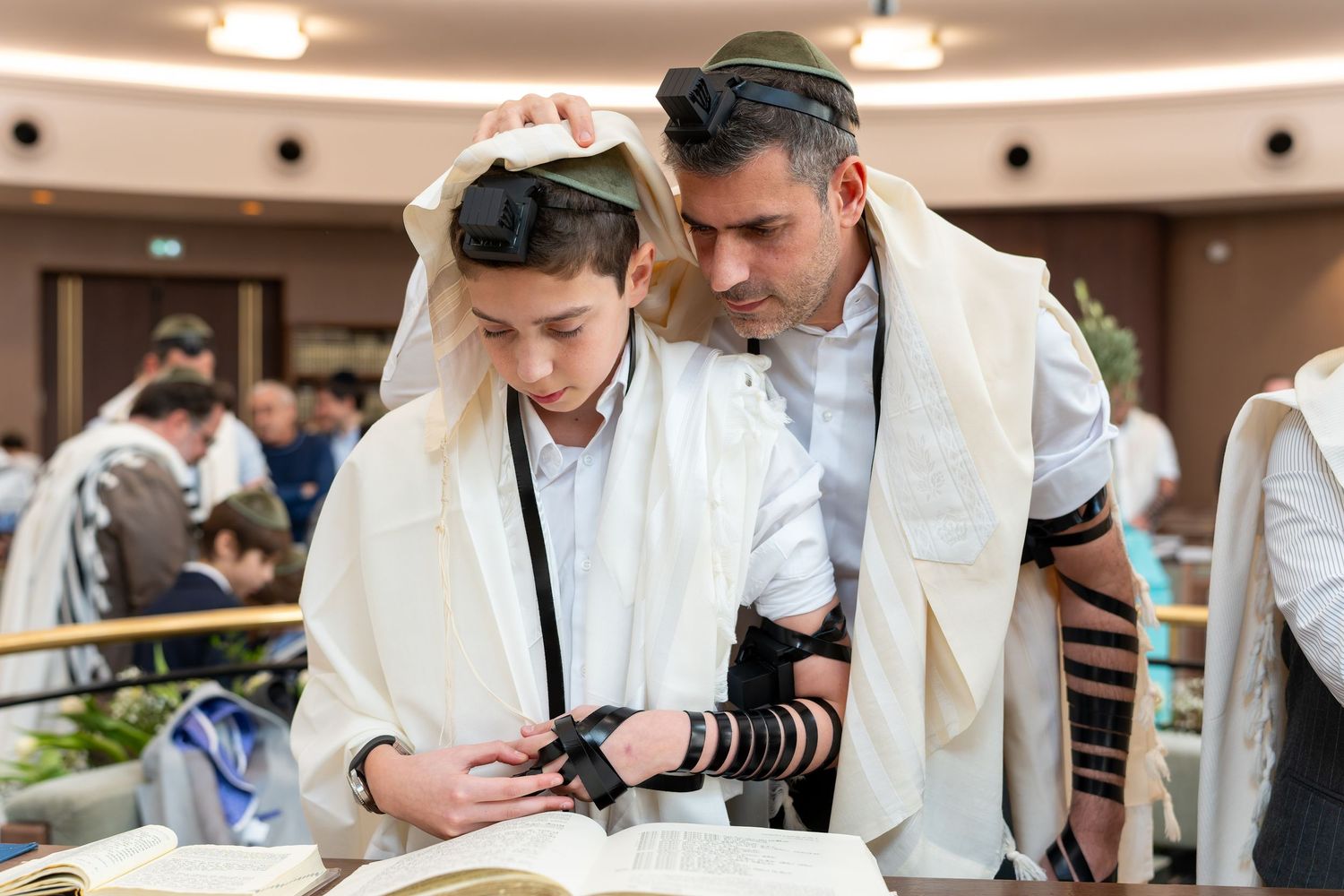 Father helps son wrap tefillin during Bar Mitzvah ceremony in synagogue.