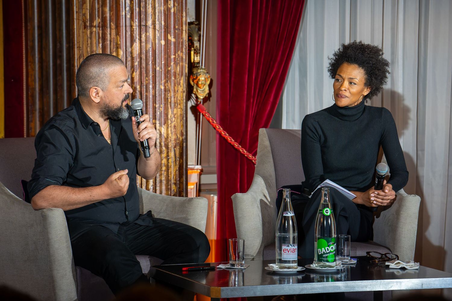 Two people engaged in a panel discussion, one speaking into a microphone at an elegant venue.