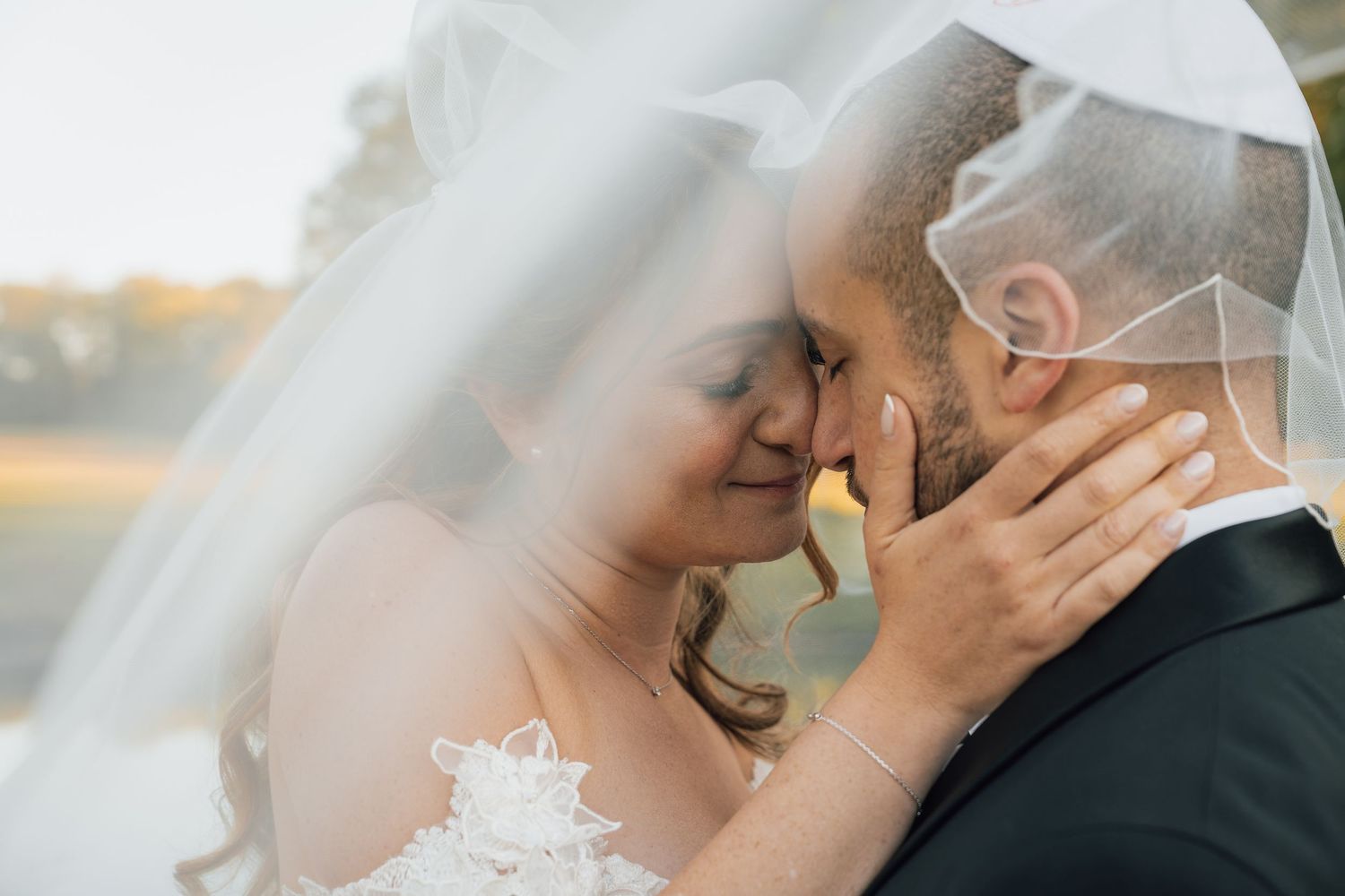 Bride and groom share tender veil-covered moment, touching foreheads in romantic outdoor wedding portrait.