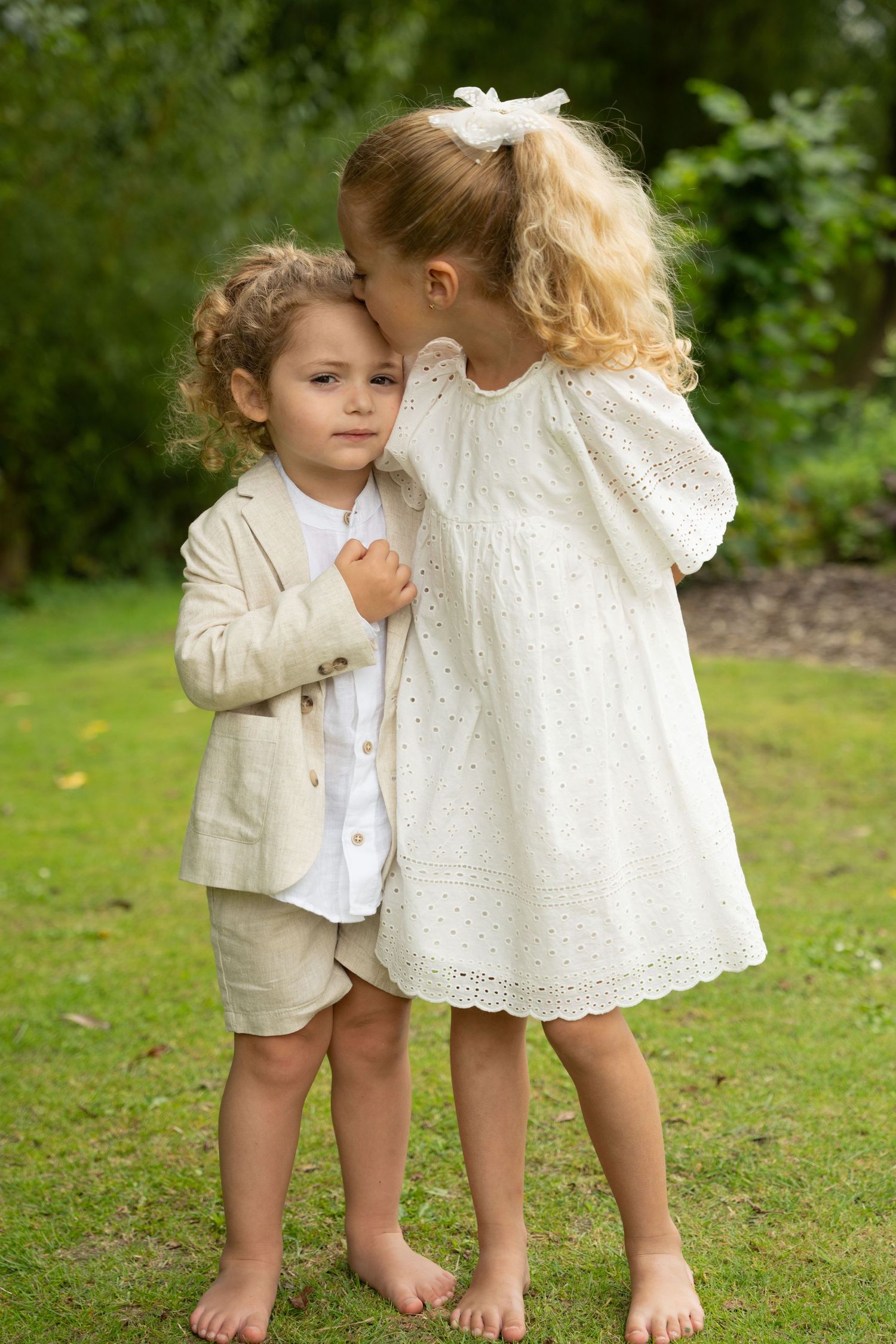 Two young children in garden, older blonde girl whispering to younger curly-haired child.