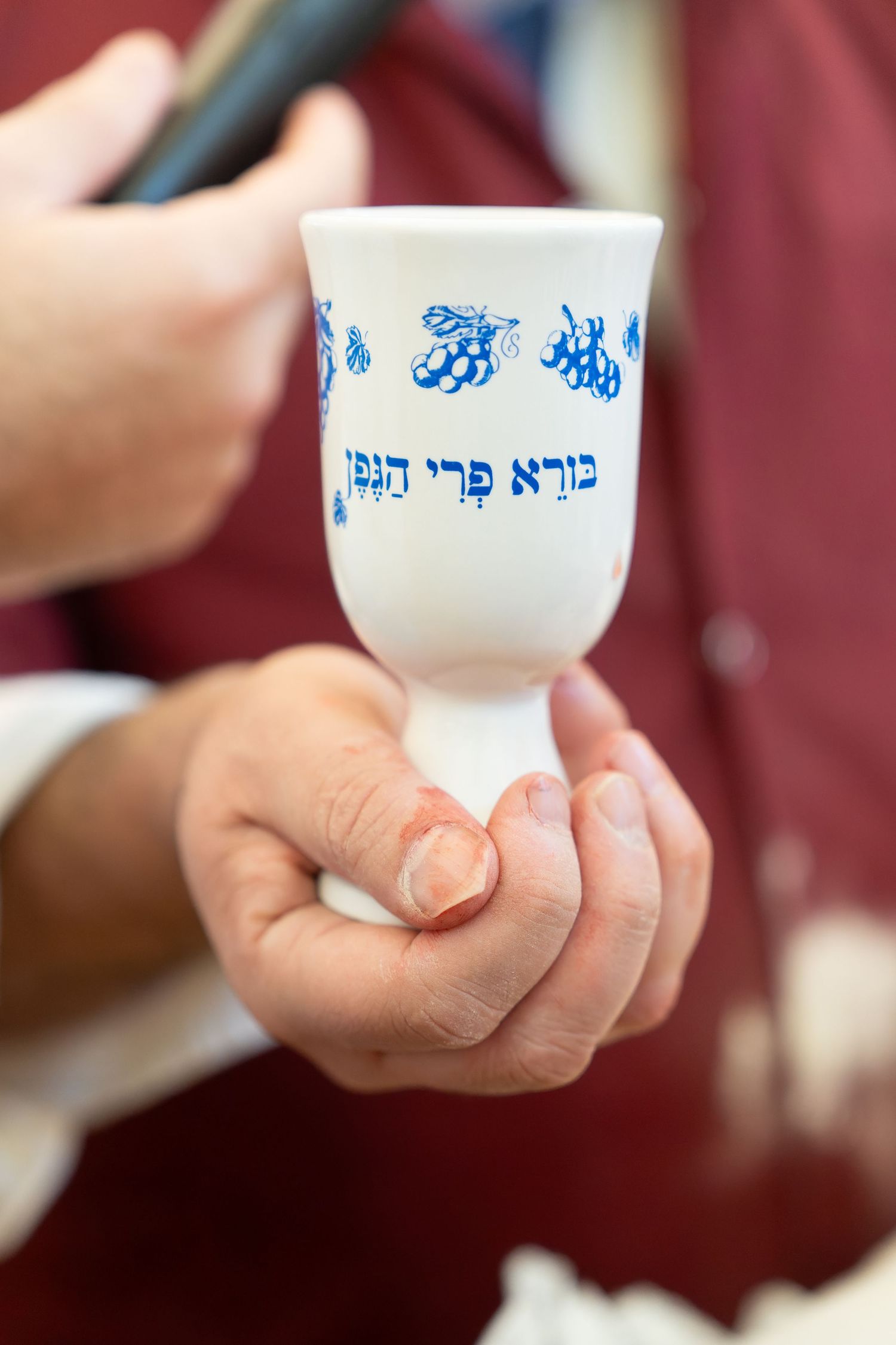 Hands holding a white Kiddush cup with blue Hebrew text and grape motifs during a Jewish ceremony.