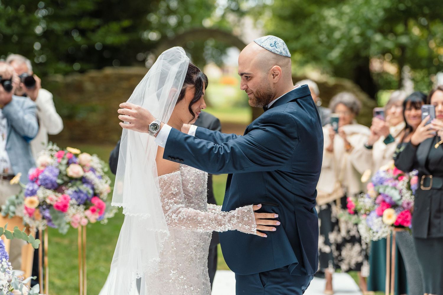 Groom lifting bride's veil for a tender moment at a colorful outdoor Jewish wedding ceremony.