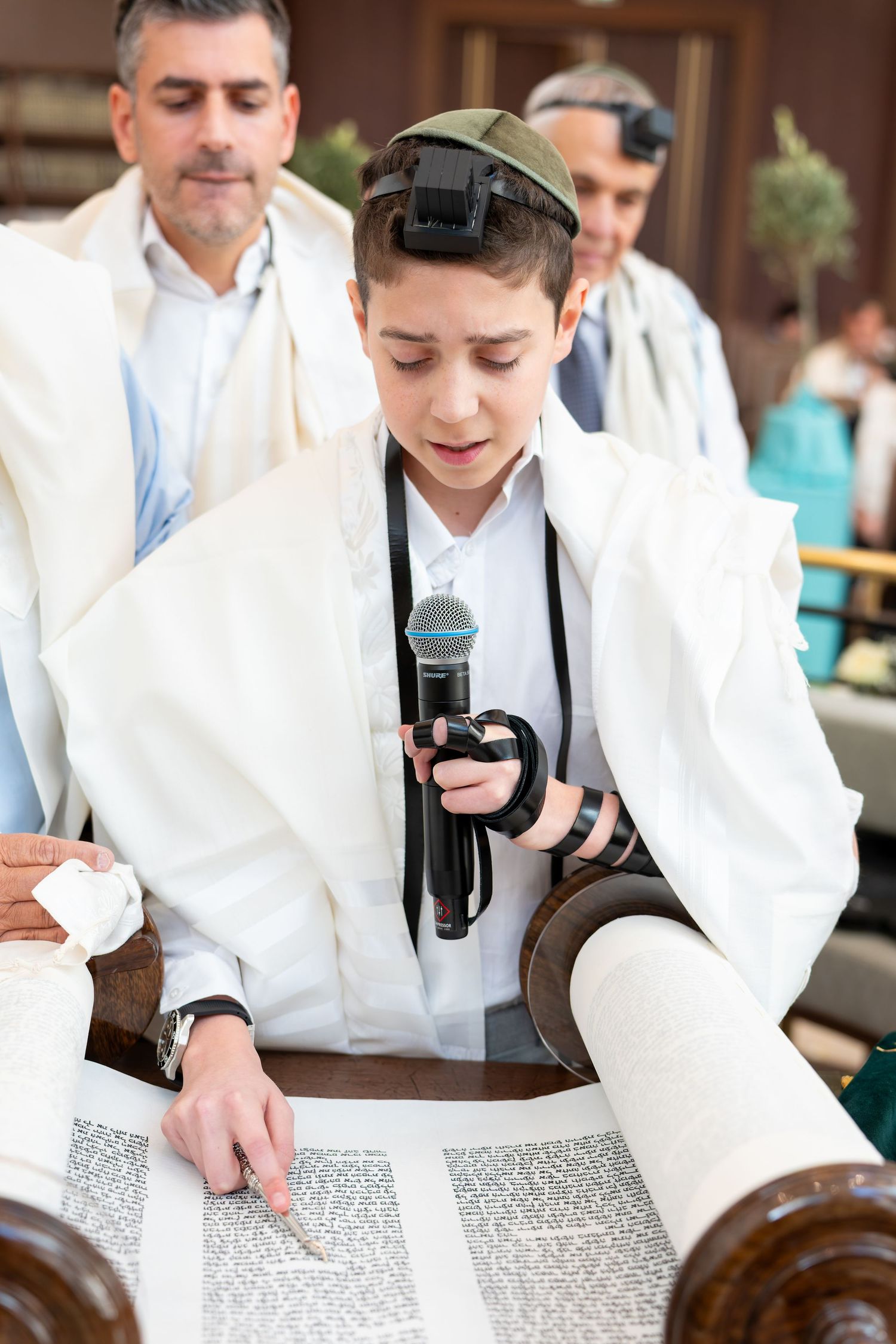 Boy reads Torah scroll at Bar Mitzvah ceremony wearing tallit and tefillin while holding microphone.