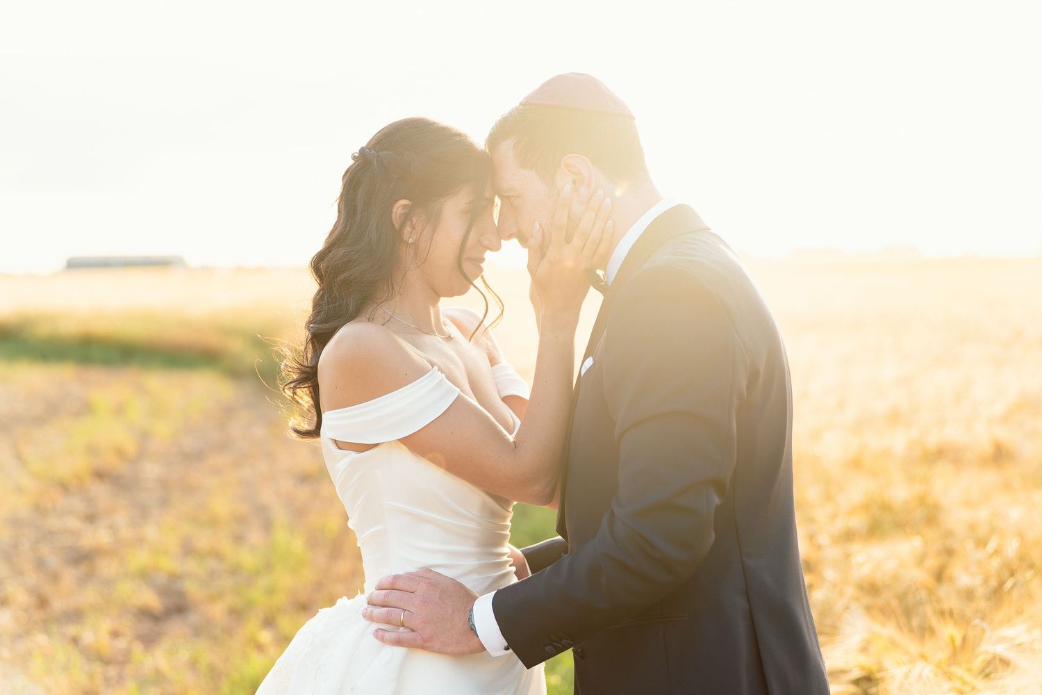 Bride and groom touching foreheads tenderly in a golden sunlit wheat field on their wedding day.