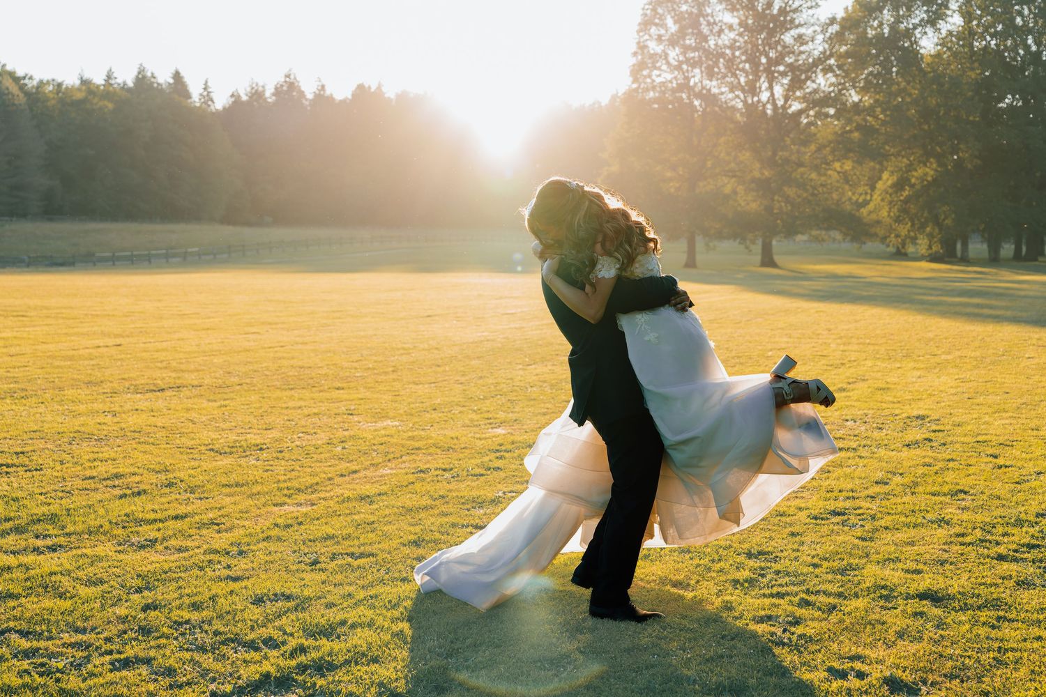 Groom lifts bride in white dress during golden hour sunset wedding photo in lush green park.
