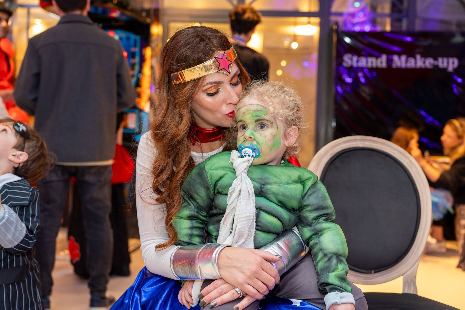 Woman dressed as Wonder Woman holding a baby with green face paint at a costume party.