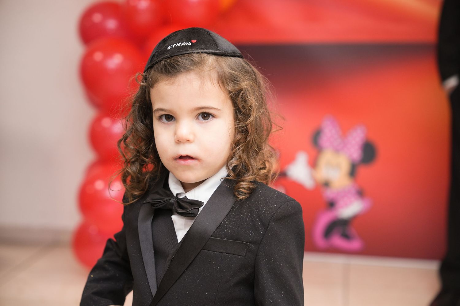 Young child in black tuxedo and kippah posing at a Minnie Mouse themed birthday party.