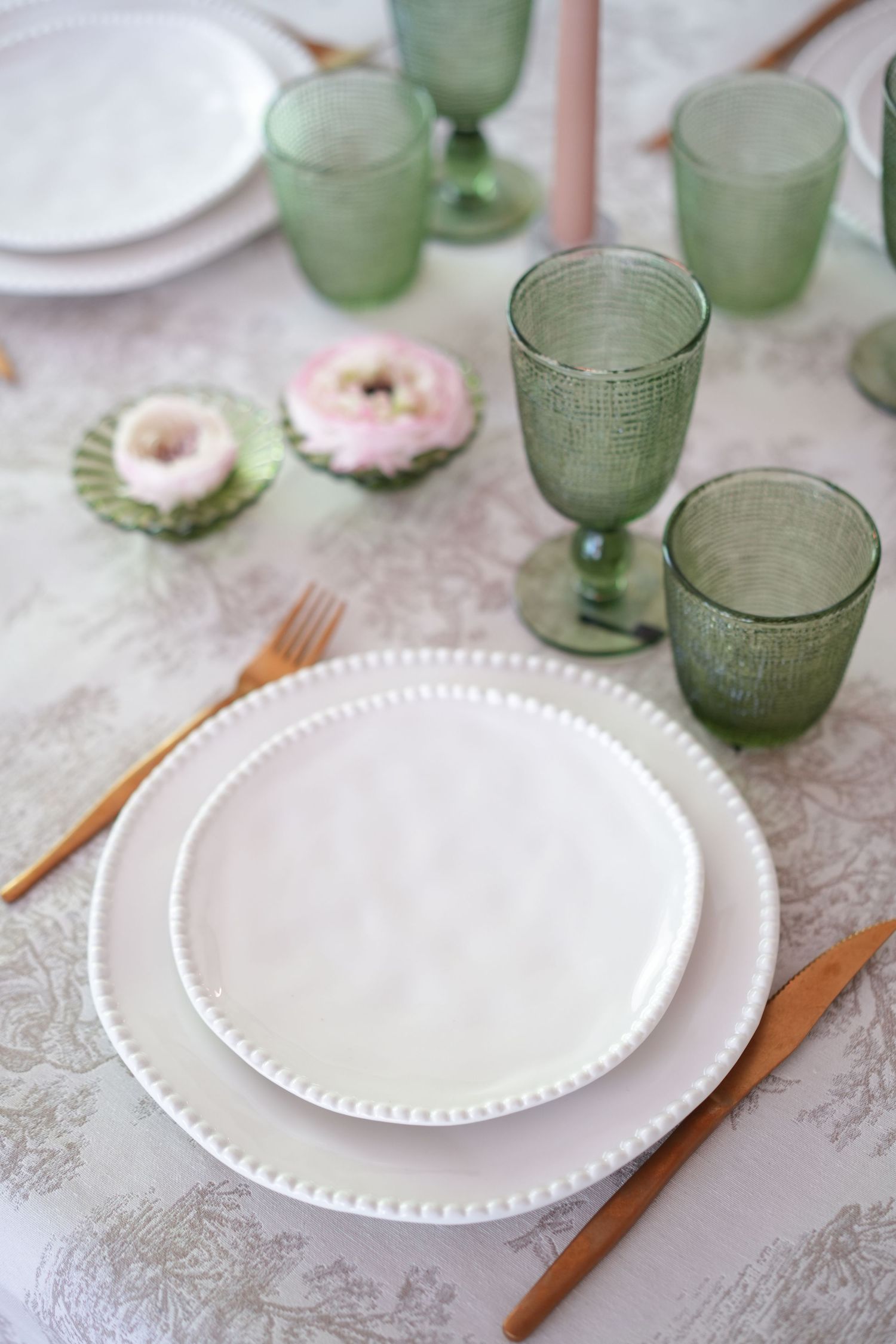 Elegant table setting with white beaded plates, gold cutlery, green glassware, and pink floral accents on a floral tablecloth.