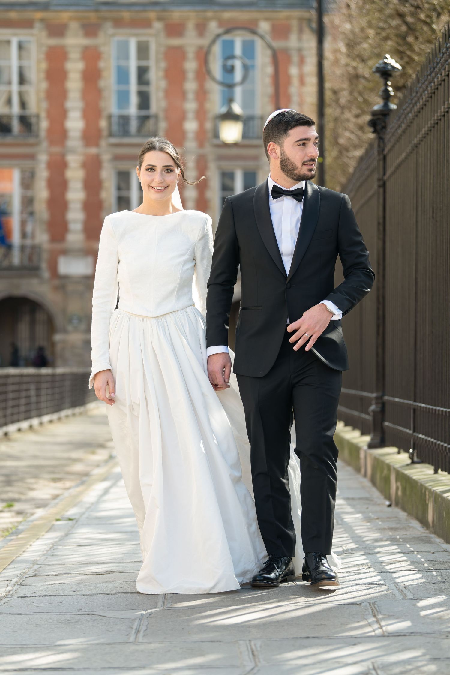Stylish wedding couple walking on city cobblestones, bride in minimalist white gown, groom in classic black tuxedo.