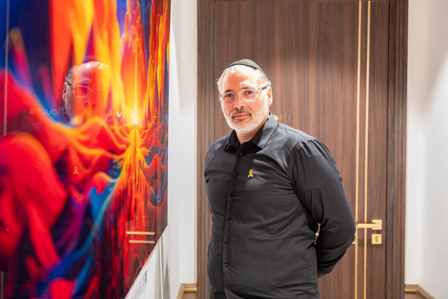 Man wearing kippah stands beside vibrant colorful artwork in modern interior setting.