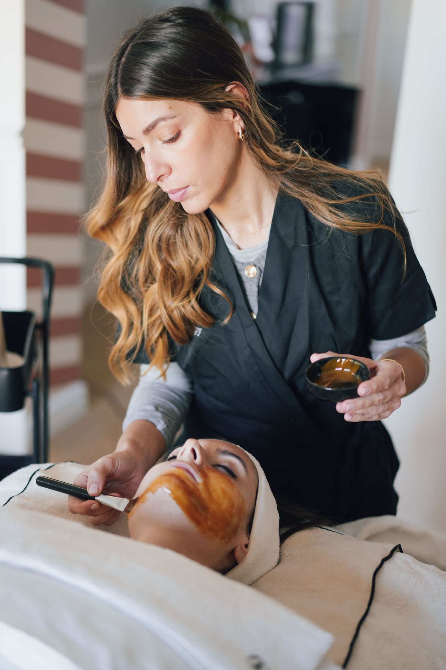 Esthetician applying brown facial mask to client using brush during professional skincare treatment.
