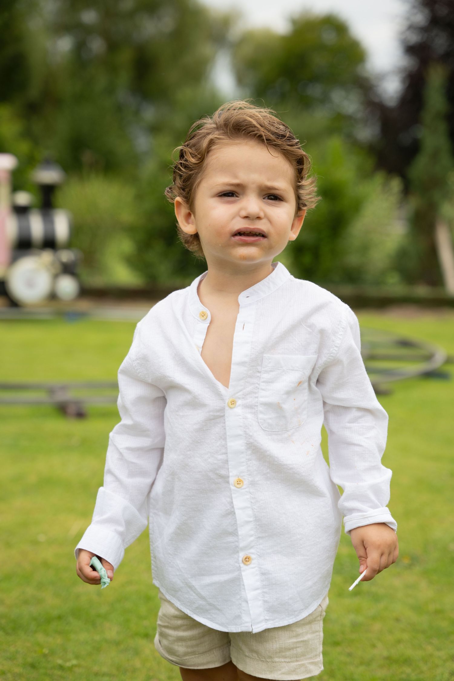 Upset toddler boy in white linen shirt standing on green grass outdoors.