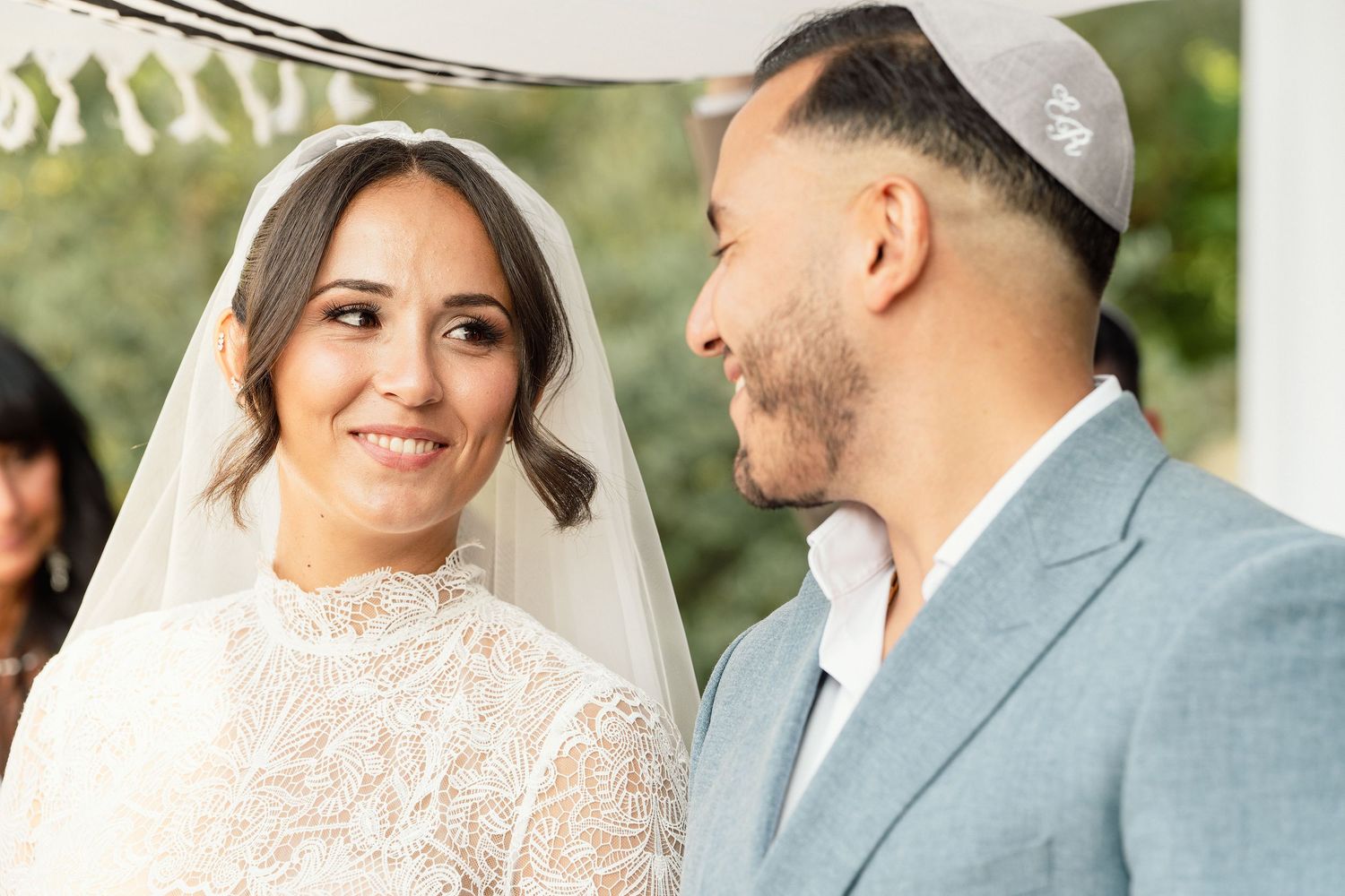 Bride in lace wedding dress and groom in gray suit exchanging vows at an outdoor Jewish ceremony.
