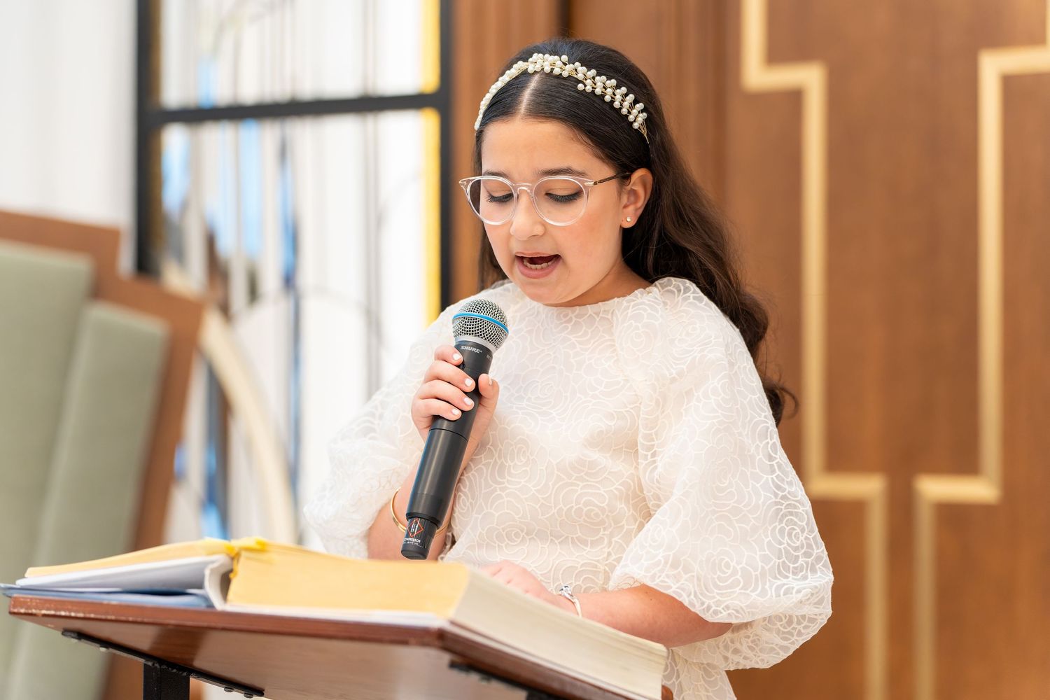 Young girl in white lace dress with pearl headband reading from book at podium with microphone.
