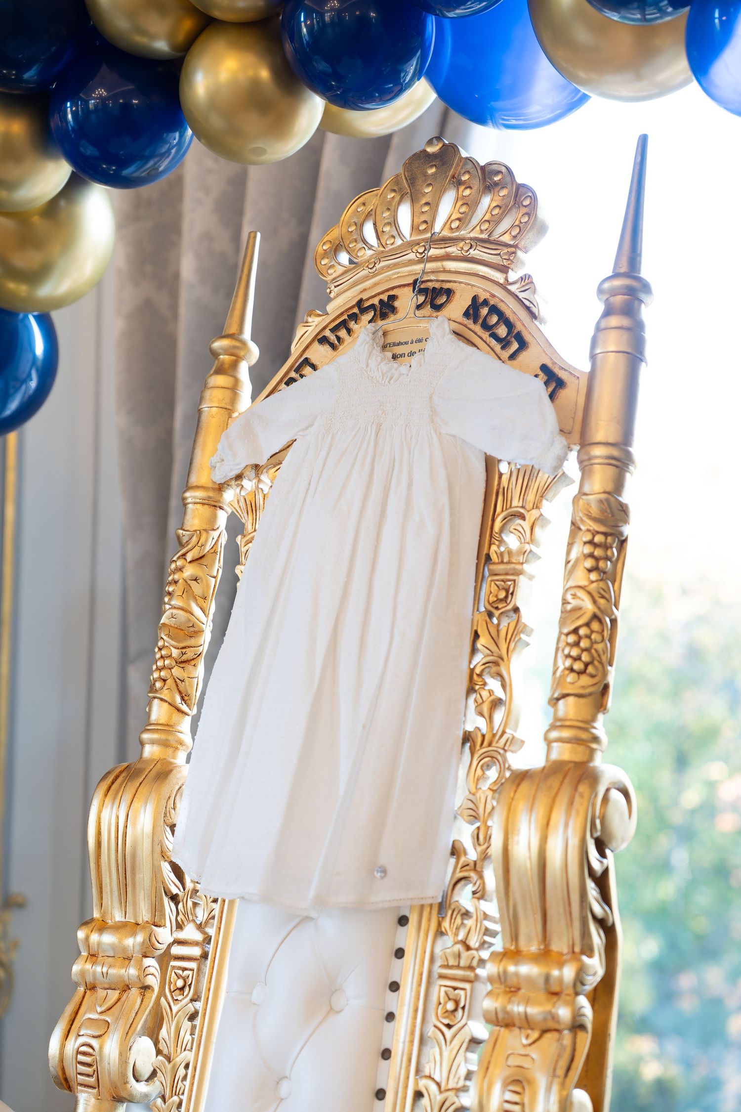 Ornate gold throne chair with white garment and Hebrew text, decorated with blue and gold balloons for a Jewish celebration.