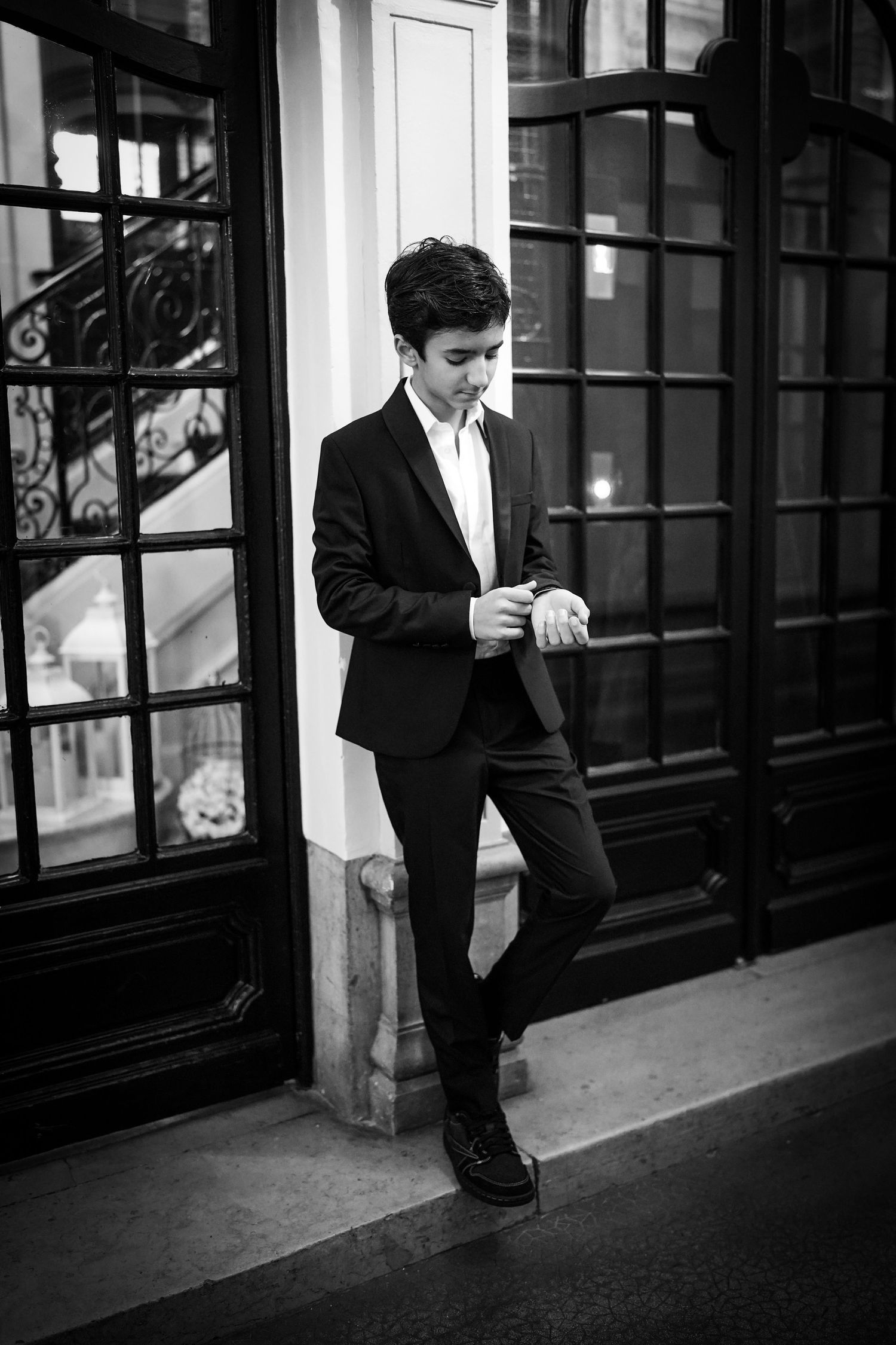 Teenage boy in black suit leans against ornate door in black and white photo.