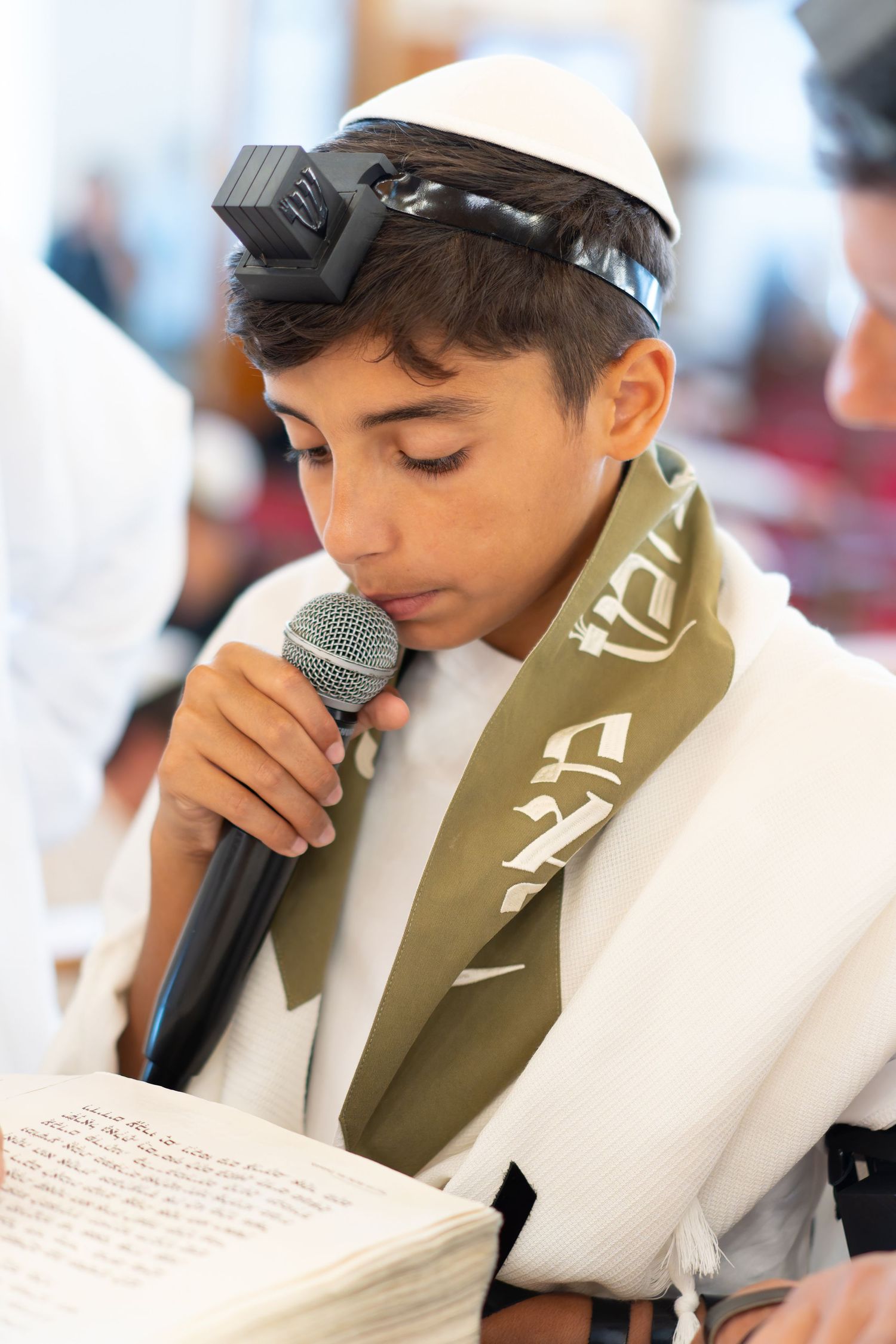 Jewish boy in white kippah and tallit reads from Torah scroll at Bar Mitzvah.