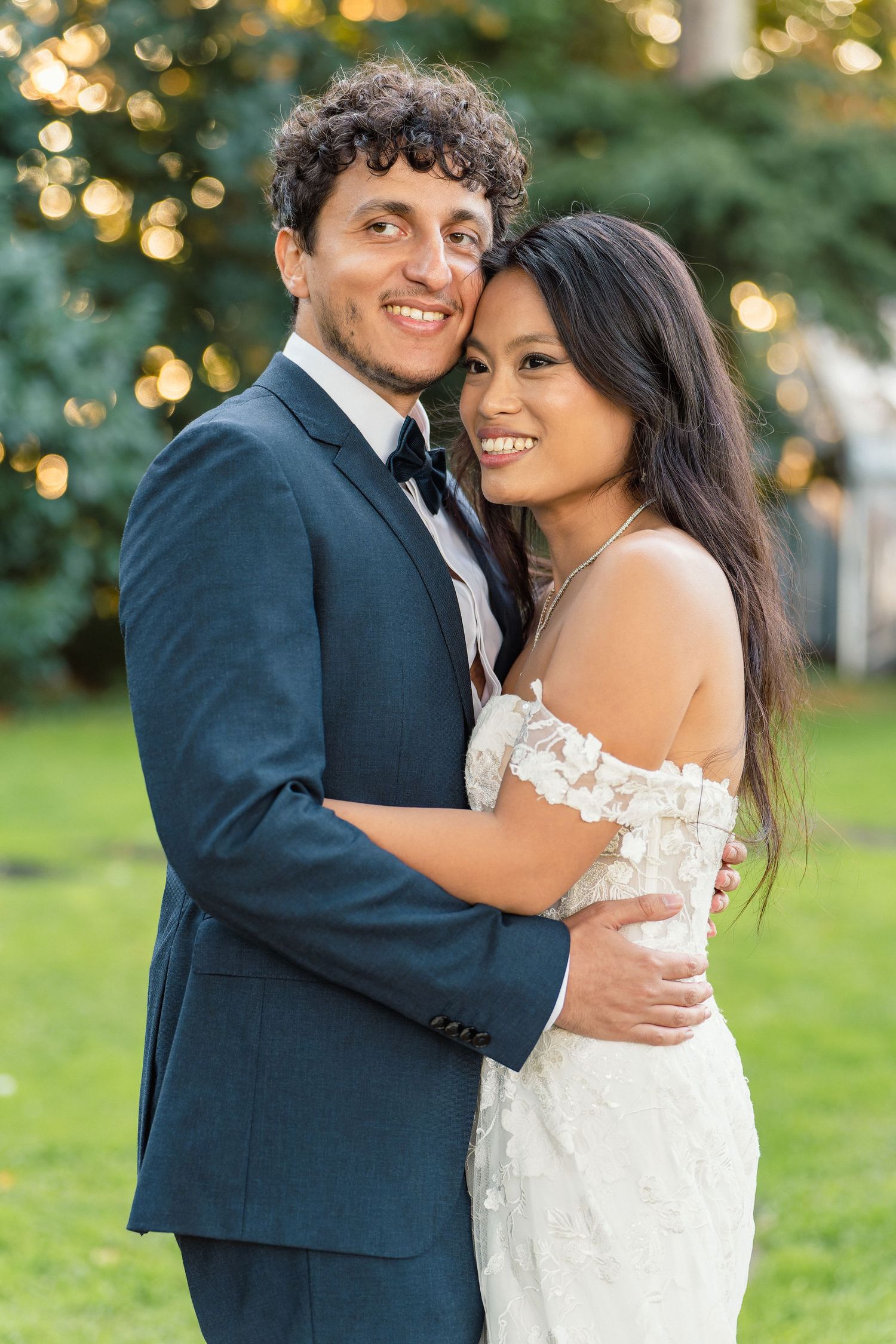 Joyful newlywed couple embracing outdoors, groom in navy suit with bow tie, bride in off-shoulder floral lace dress.