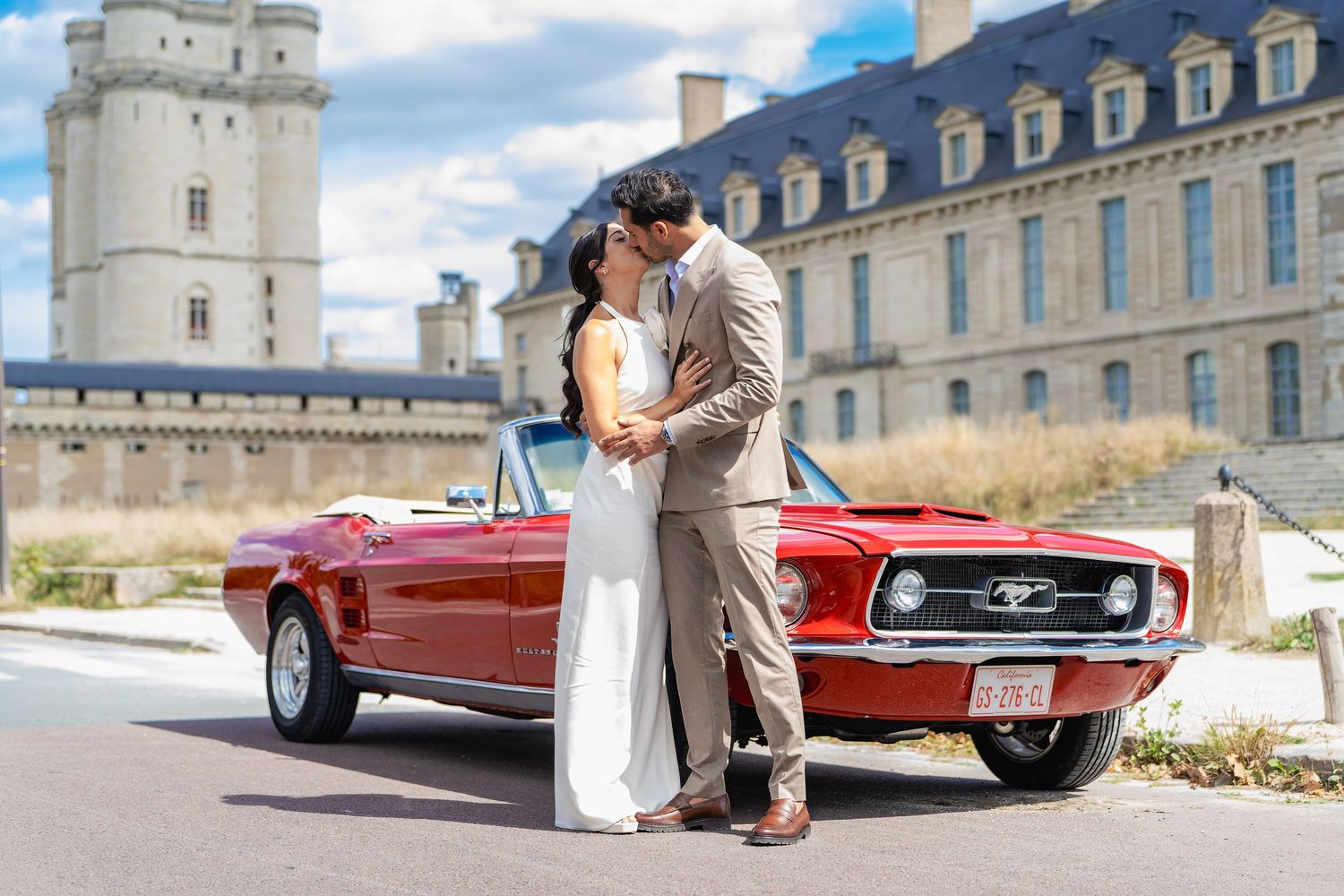 Wedding couple kissing beside a classic red Ford Mustang convertible with a historic castle in the background.