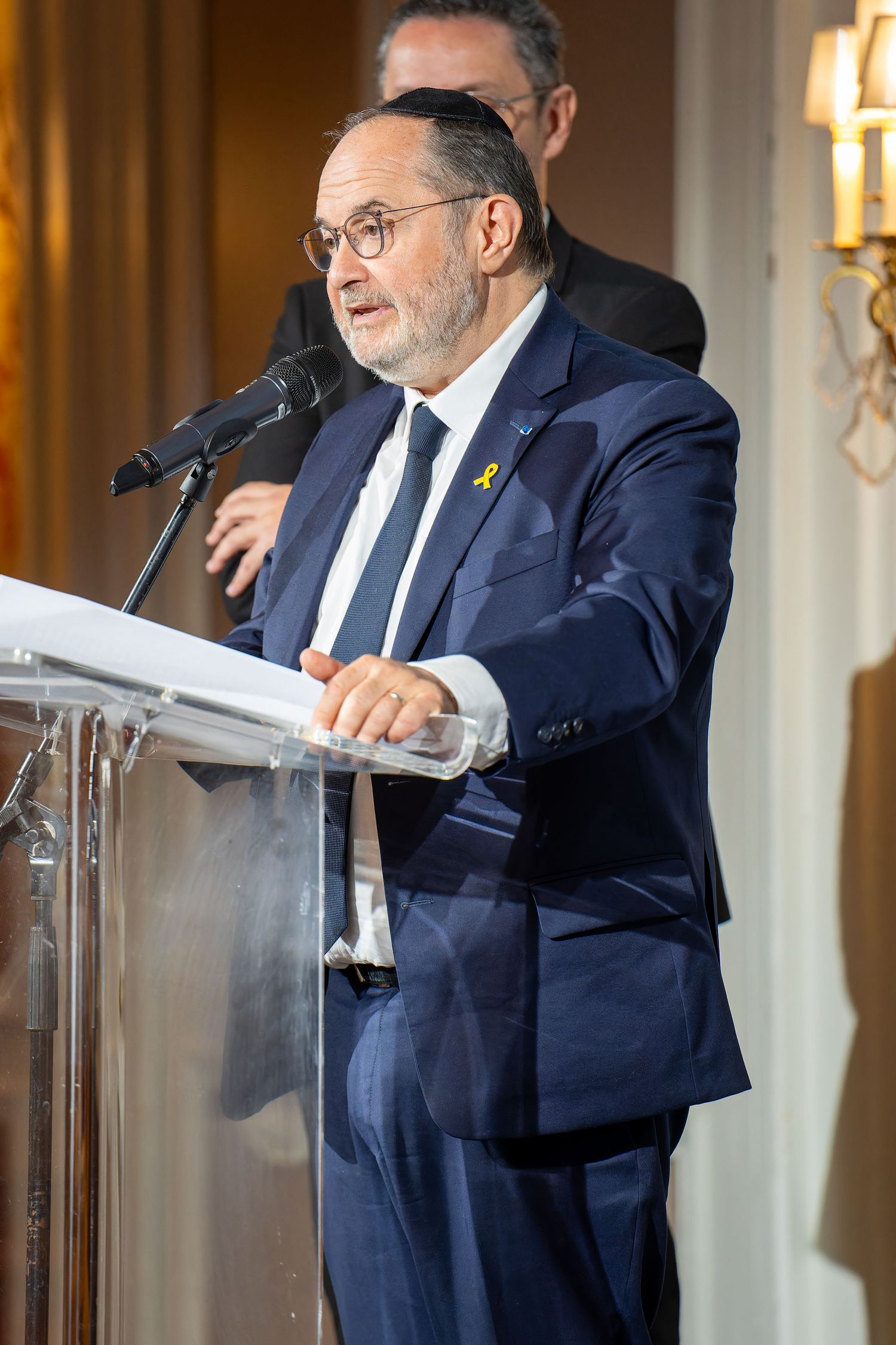 Man in navy suit with yellow ribbon speaks at podium during formal Jewish philanthropy event.